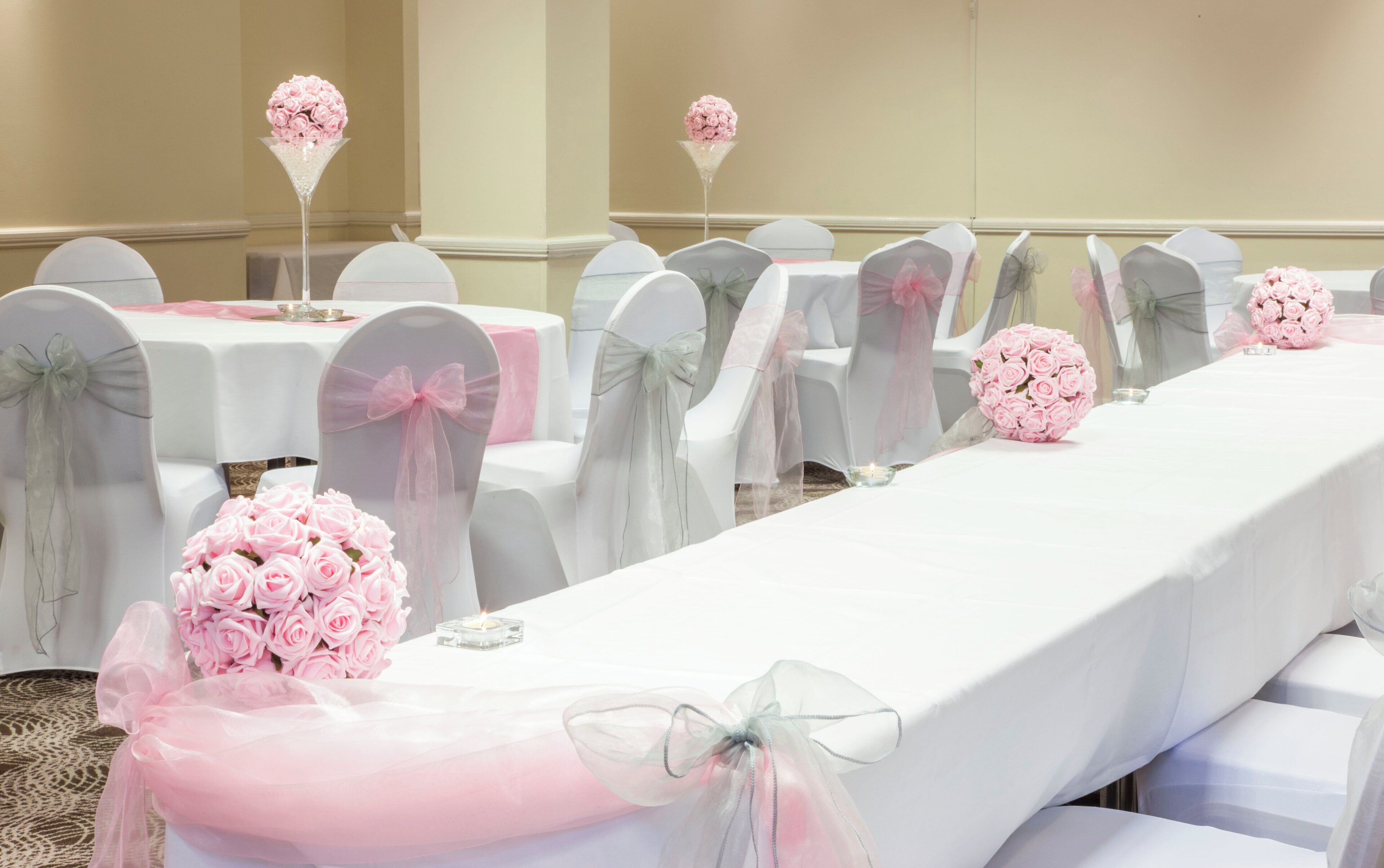 A banquet hall with rows of white tables, each with a pink flower centerpiece and a candle.