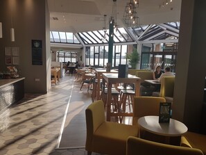 A spacious indoor area with tables and chairs, a counter, and a large window allowing natural light. - Holiday Inn Leamington Spa - Warwick by IHG (Warwick)