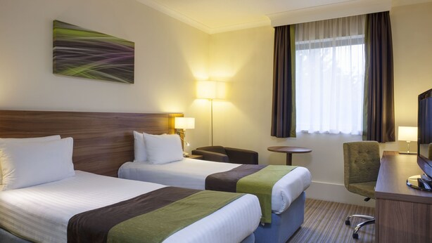 A hotel room with two beds, a desk, a chair, a lamp, and a television. - Holiday Inn Leamington Spa - Warwick by IHG (Warwick)