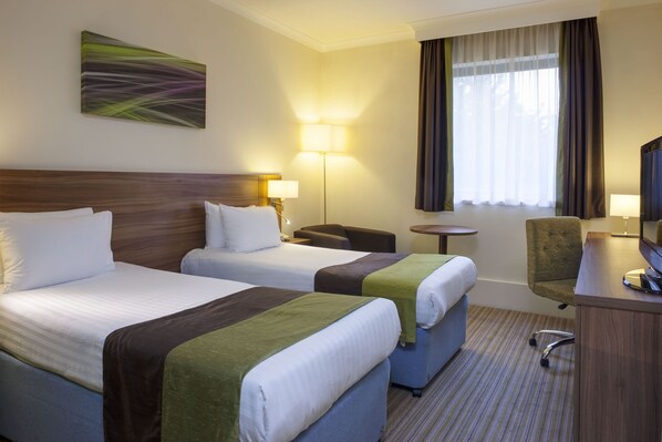 A hotel room with two beds, a desk, a chair, a lamp, and a television.