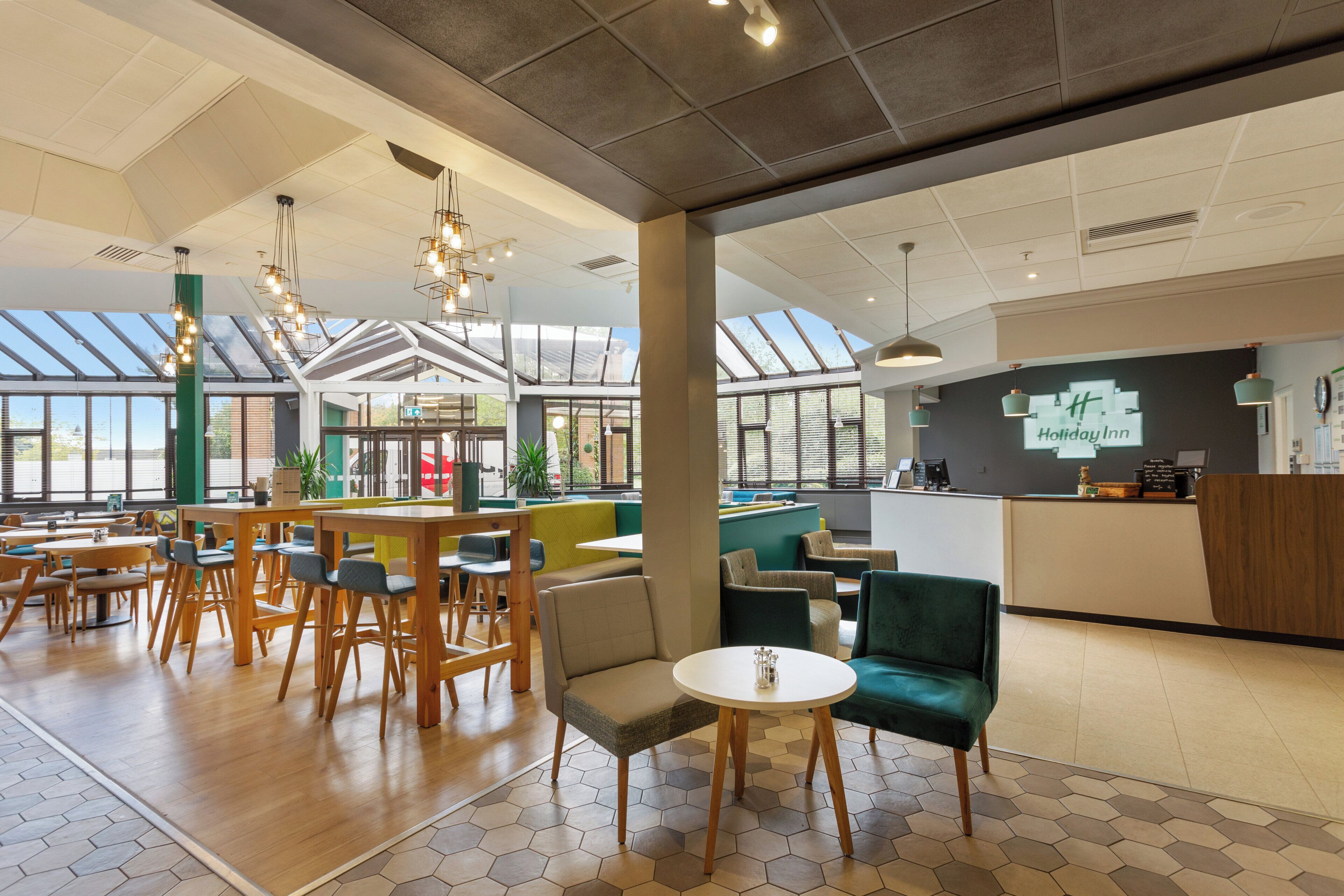 A spacious indoor area with high ceilings, wooden tables, and chairs, featuring a reception desk and a large window allowing natural light.