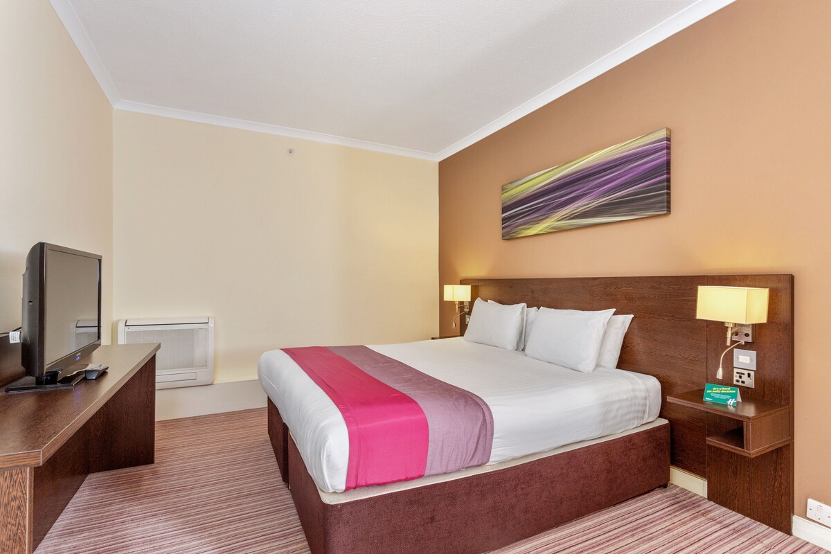 A hotel room with a large bed, a desk with a television, a wall-mounted air conditioner, and a painting above the bed.