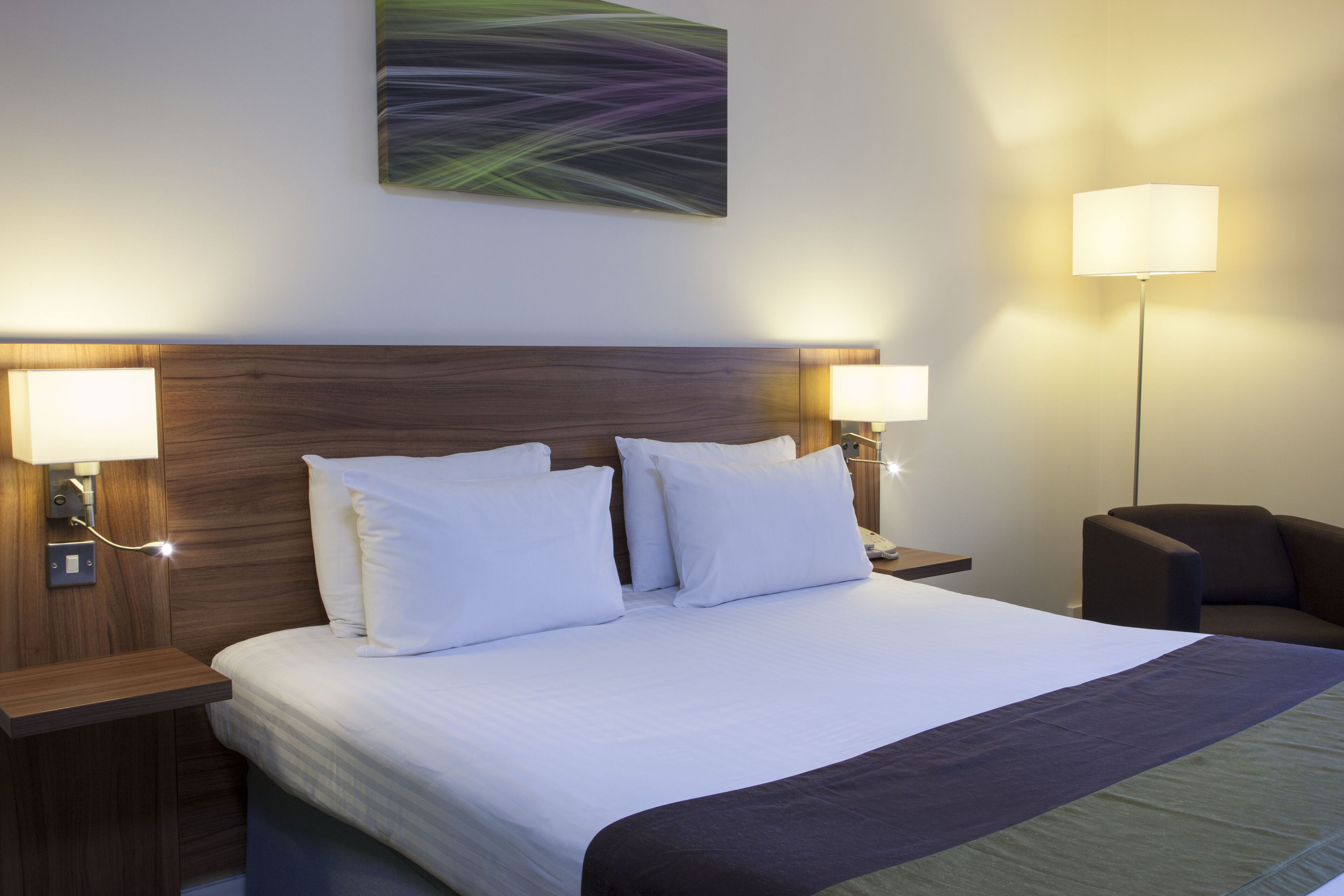 A neatly made bed with white linens and a dark-colored throw blanket, flanked by two bedside tables with lamps, a wooden headboard, and a wall-mounted abstract painting.
