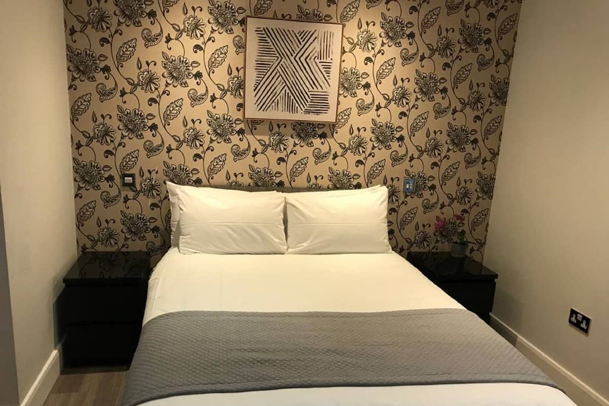 A neatly made bed with white linens and a grey blanket, positioned against a wall with a floral wallpaper pattern.