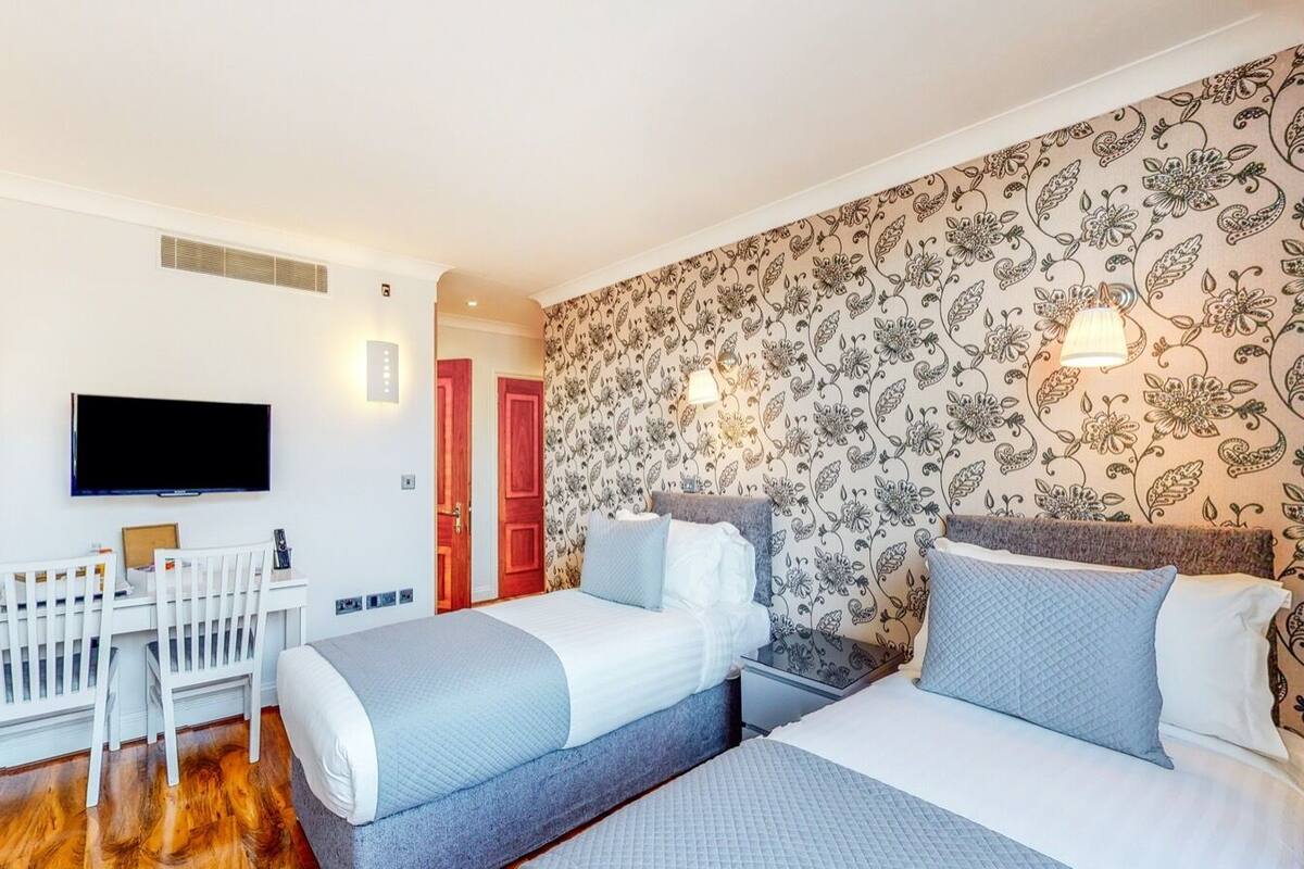 A hotel room with two beds, a desk, a TV, and a patterned wall.