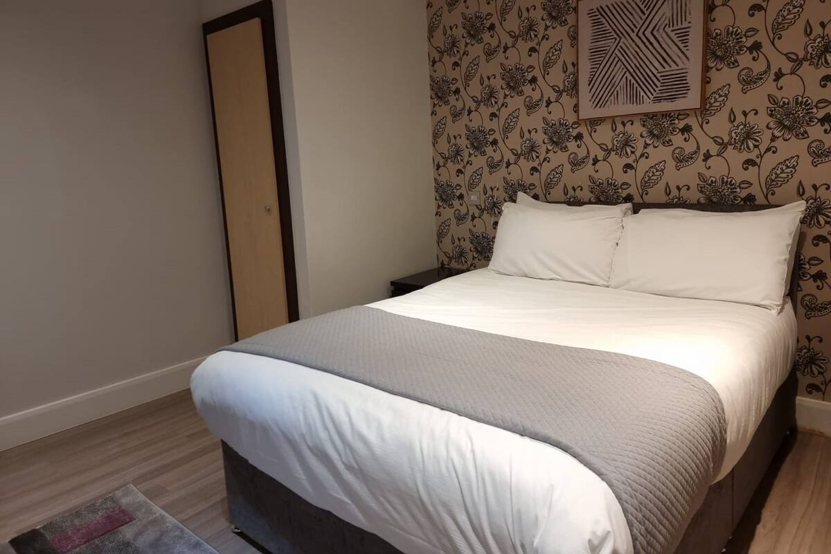 A neatly made bed with white linens and a grey blanket in a room with floral wallpaper and a geometric wall art piece.