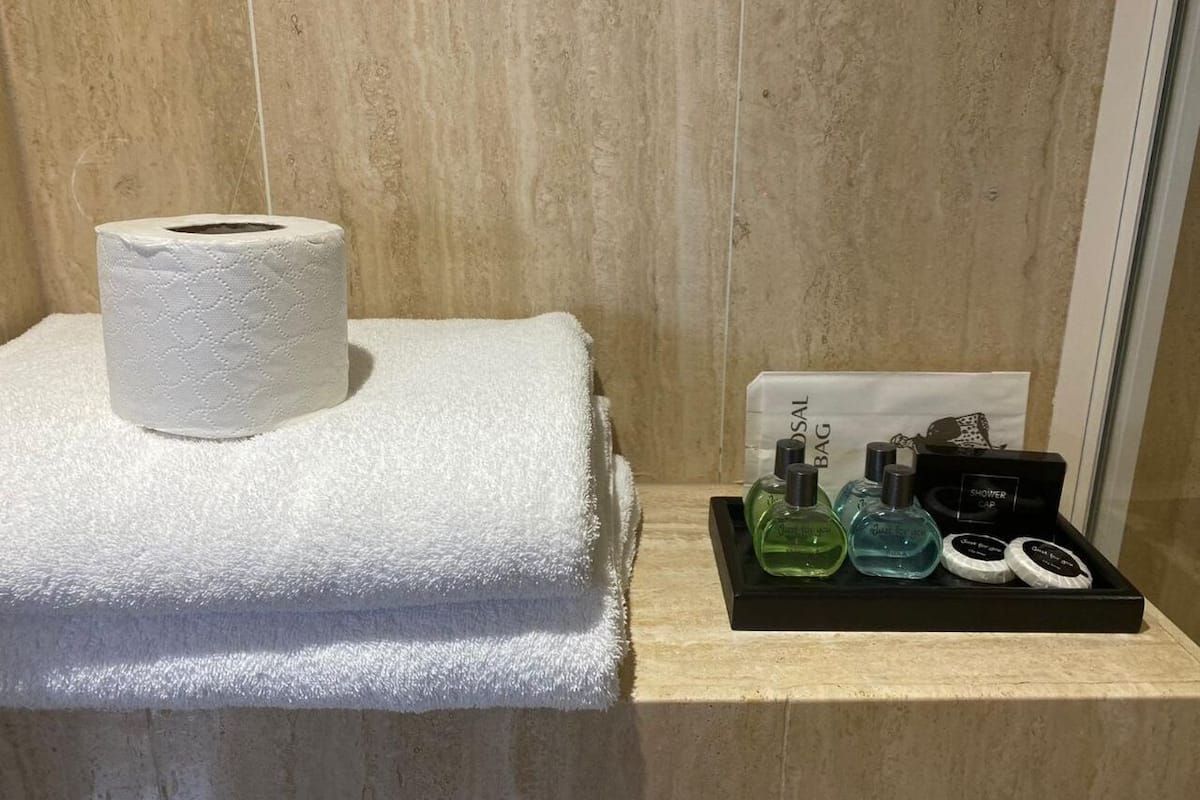 A bathroom with a white towel, toilet paper, and a tray with toiletries including a green bottle, a blue bottle, and a black container.