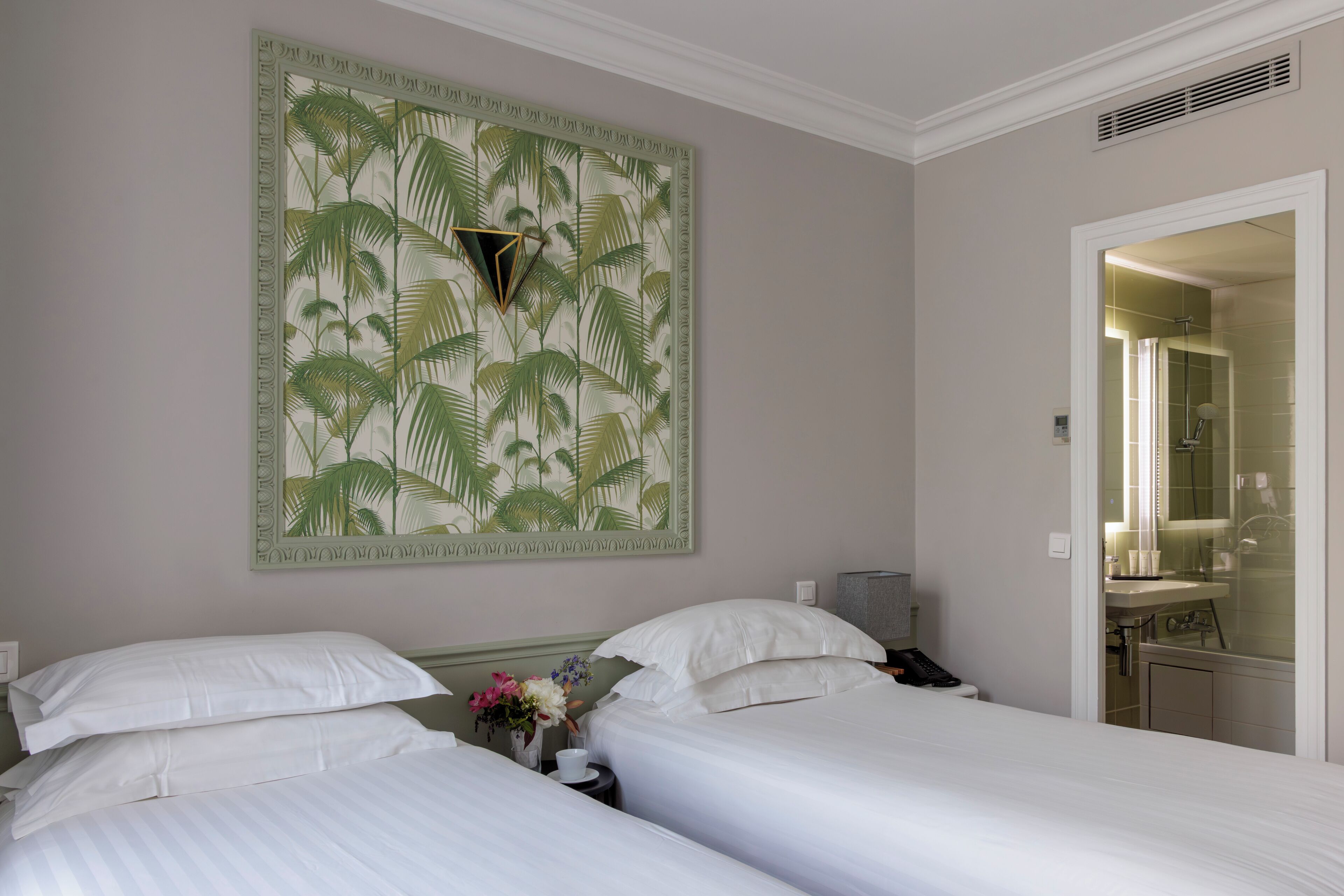 Hotelkamer met twee bedden, een groot kunstwerk van palmbomen, een badkamer met wastafel en spiegel, en een telefoon op het nachtkastje.