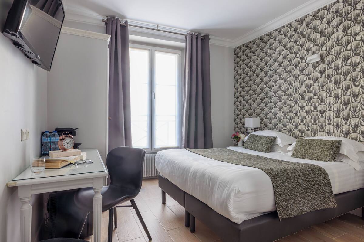 A hotel room with a bed, a desk with a chair, a TV mounted on the wall, and a window with curtains.