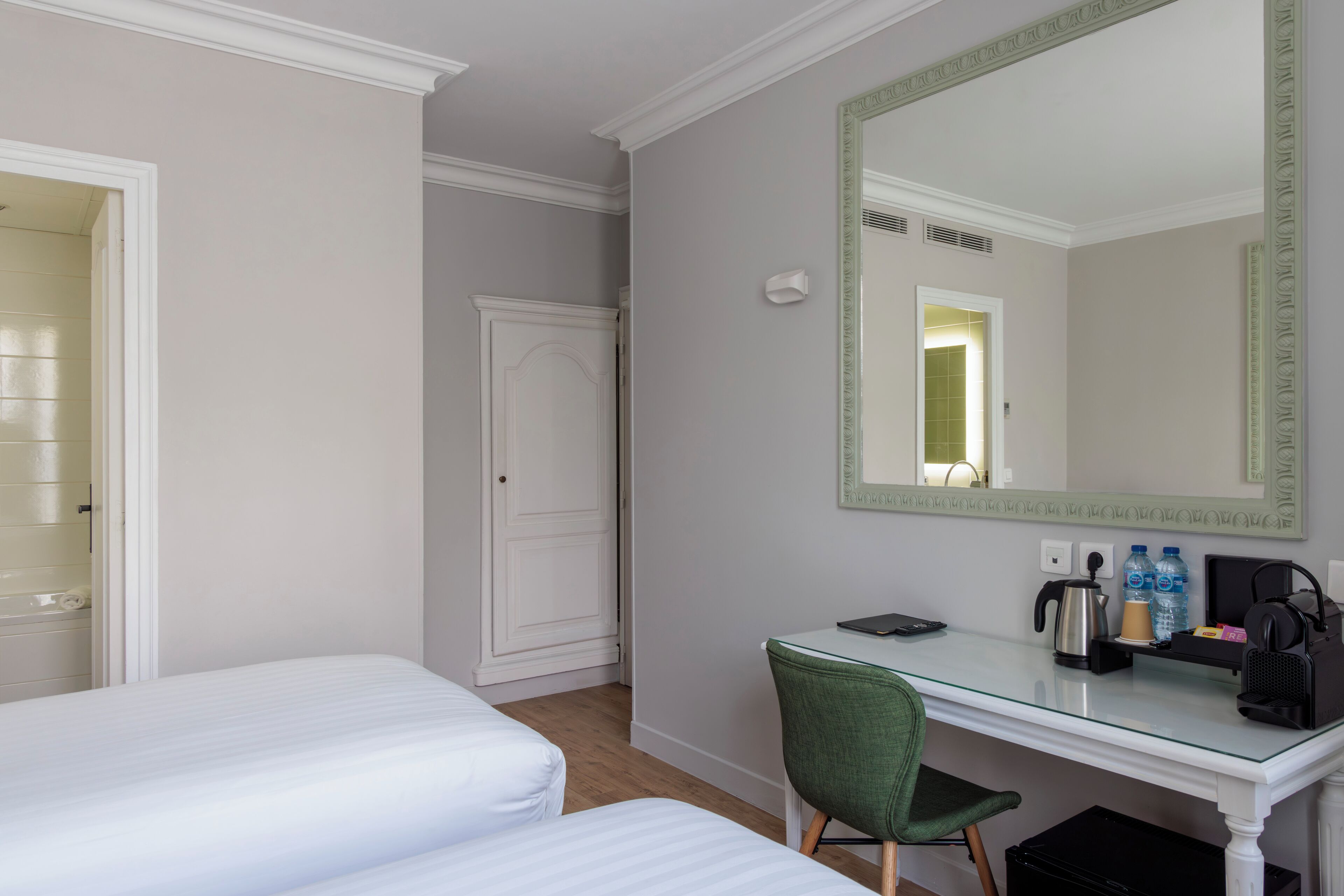 A hotel room with two beds, a desk, a chair, a mirror, a kettle, and a small trash bin.