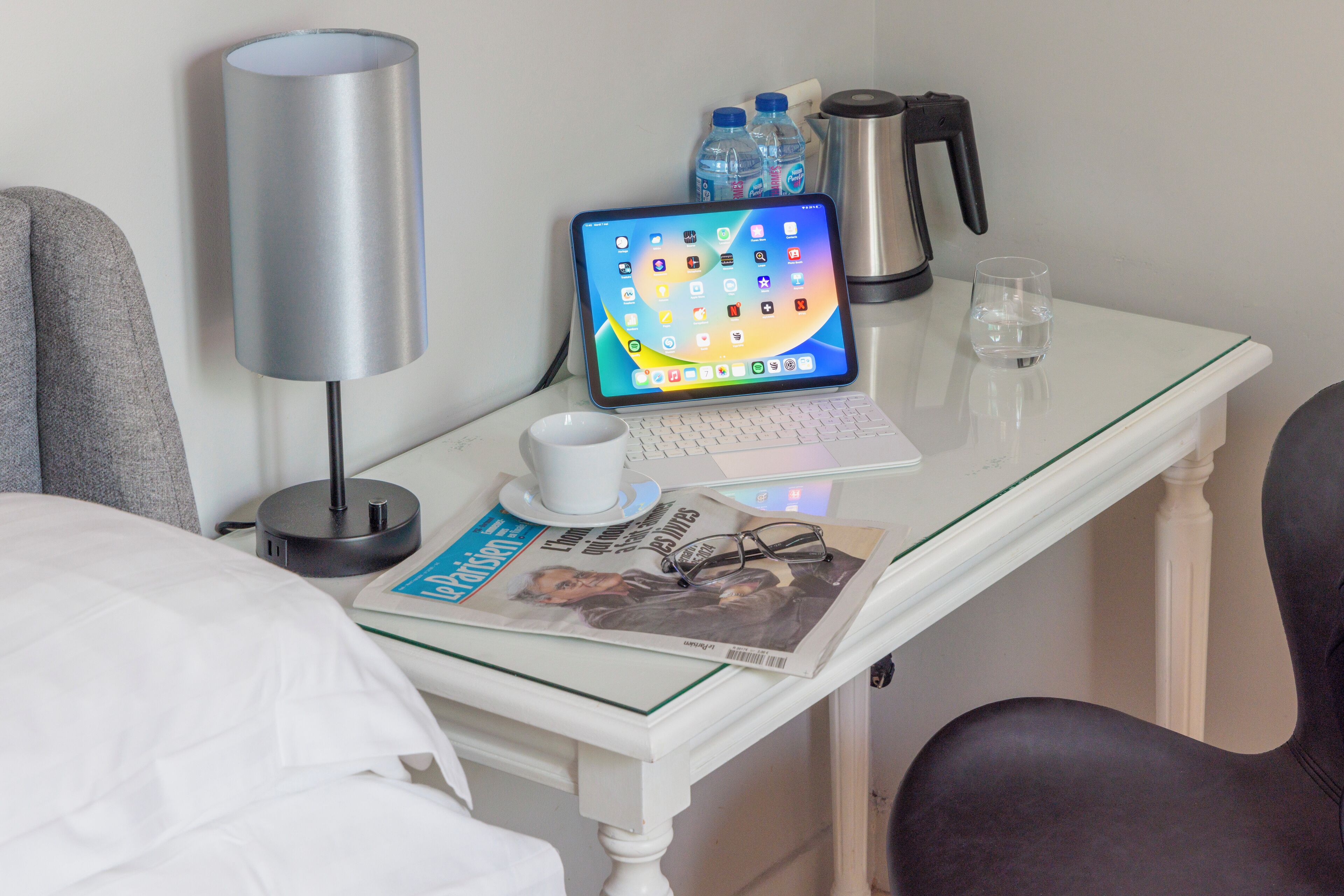 Habitación de hotel impecable con un escritorio de vidrio, una computadora portátil, una tableta, una revista, un par de gafas, una botella de agua, un termo y una lámpara.