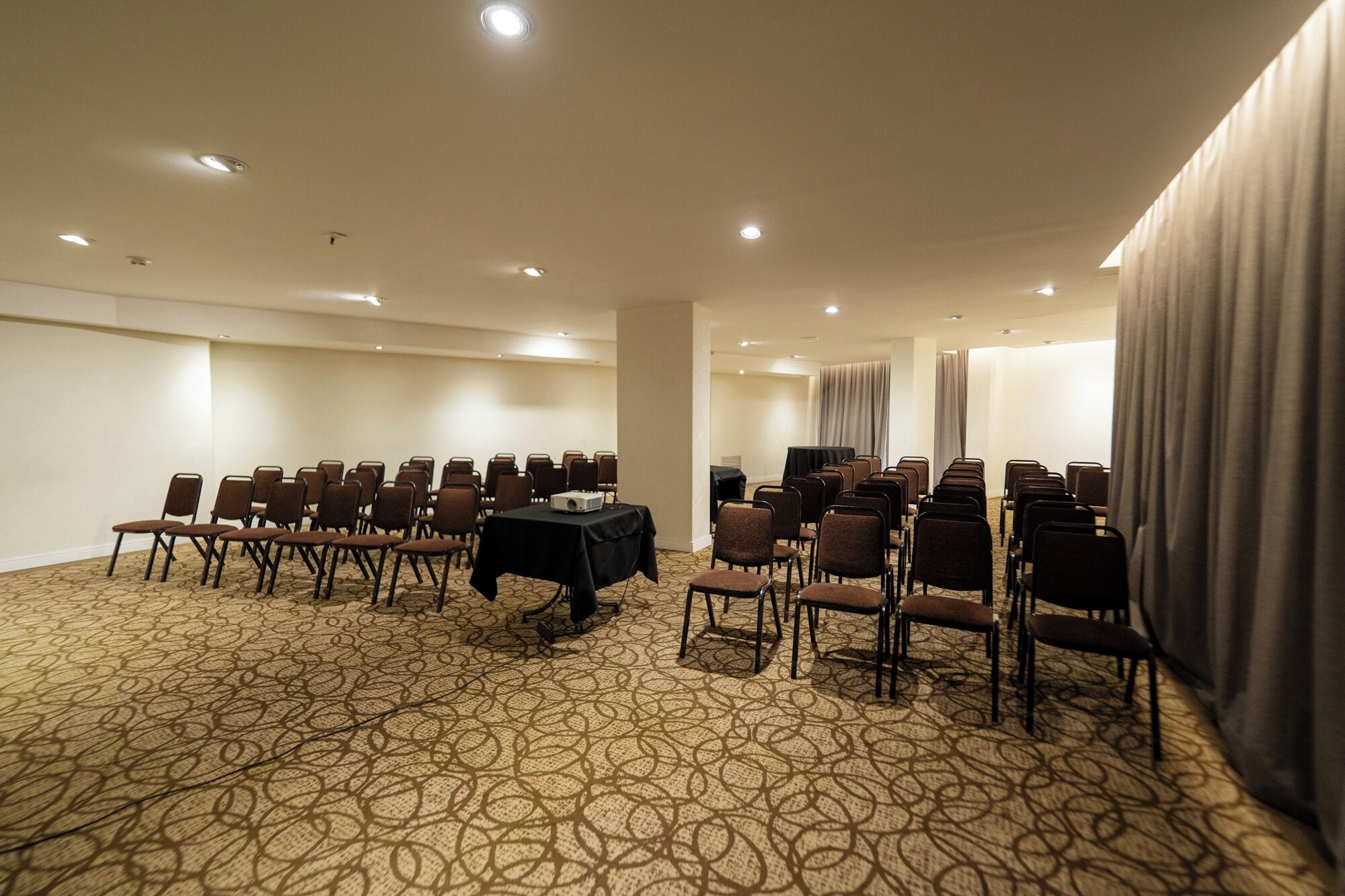 Un salón de conferencias con filas de sillas, un proyector sobre una mesa y una cortina a un lado.