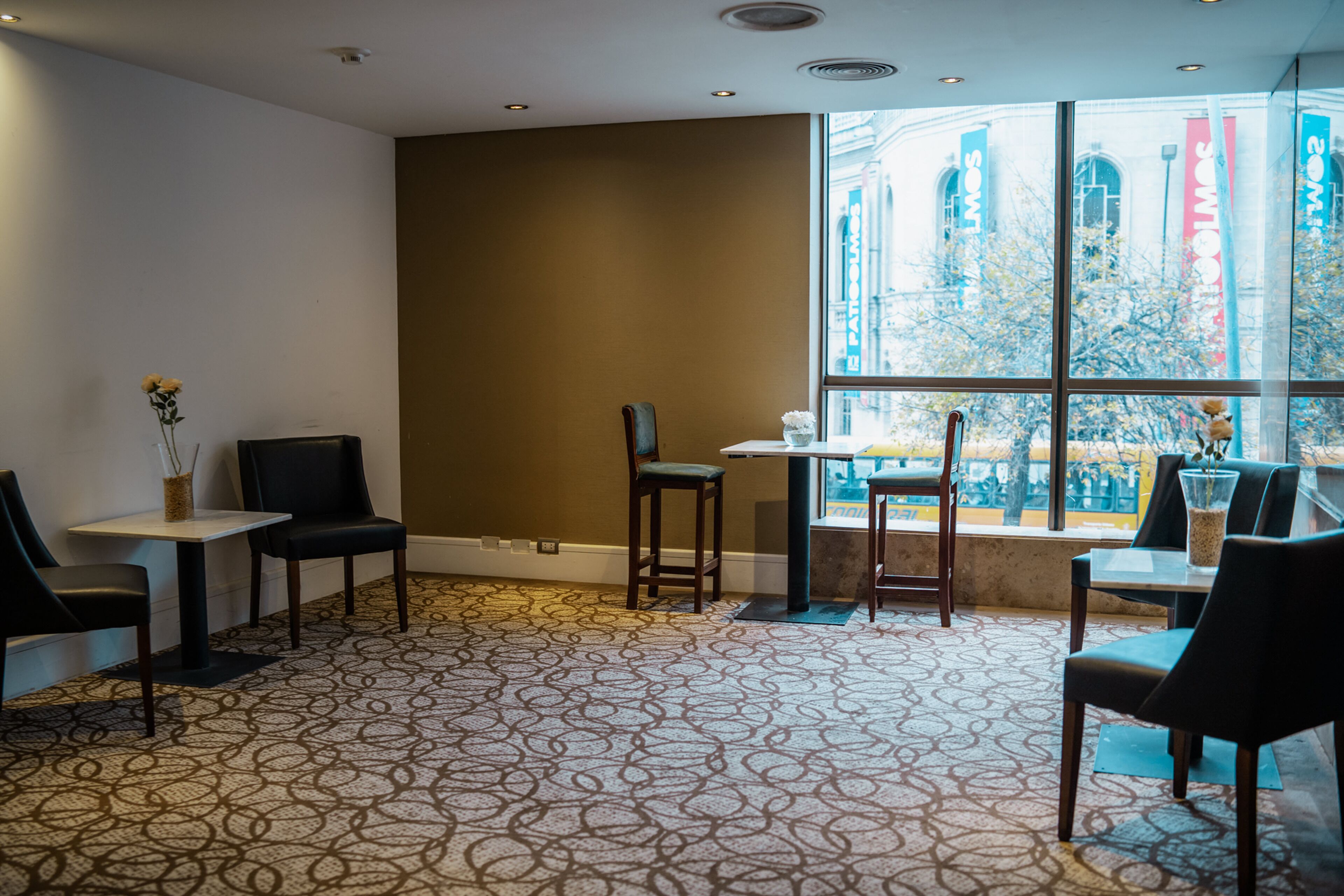 A hotel room with a patterned carpet, a large window with a view of buildings, and a seating area with chairs and tables.