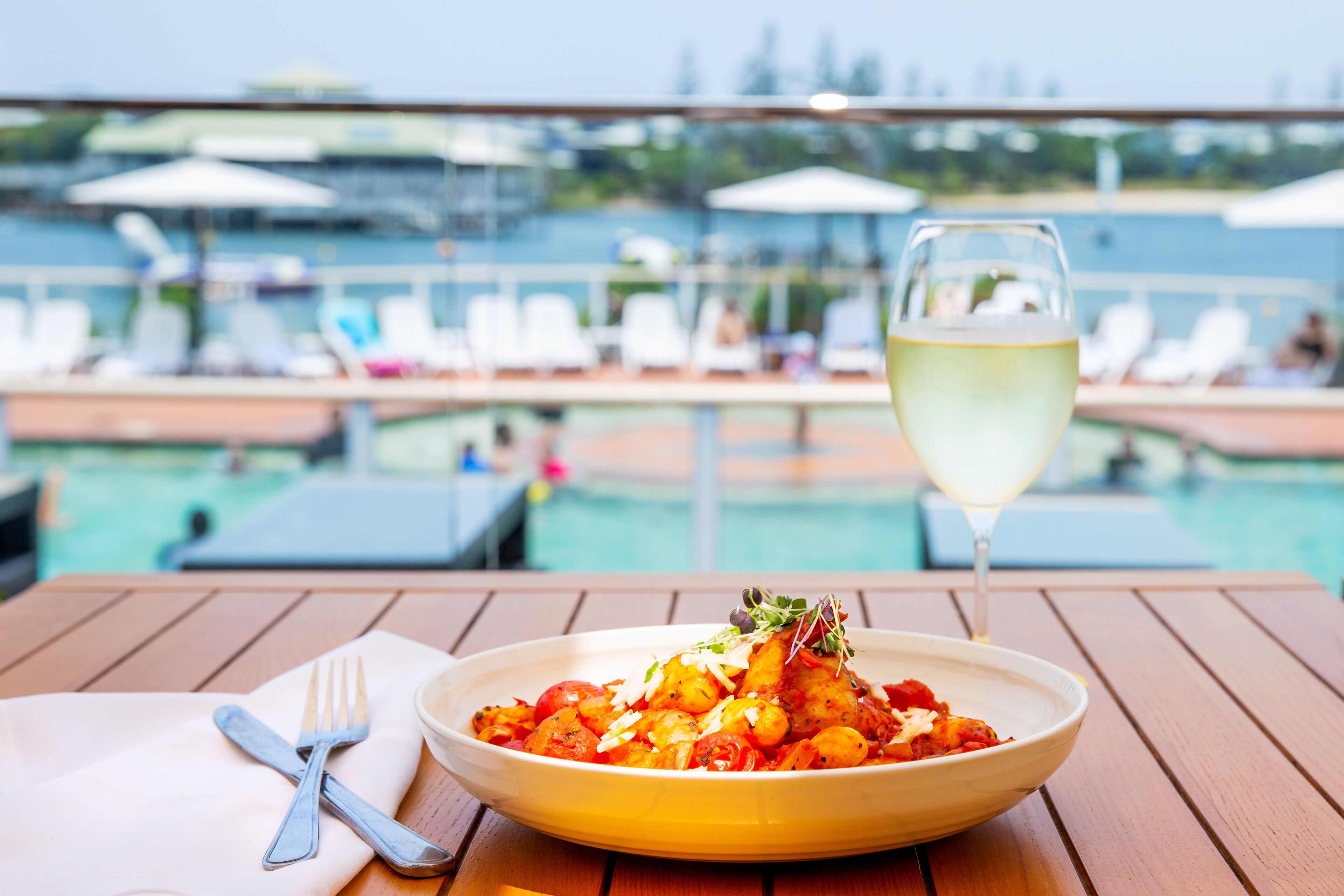 A dining table with a bowl of food, a wine glass, and cutlery on a wooden deck overlooking a pool area with lounge chairs and umbrellas.