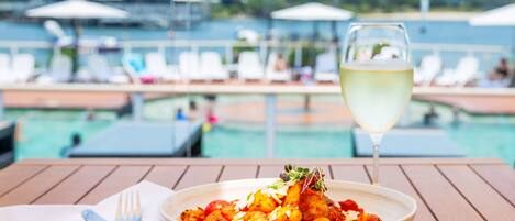 A dining table with a bowl of food, a wine glass, and cutlery on a wooden deck overlooking a pool area with lounge chairs and umbrellas.