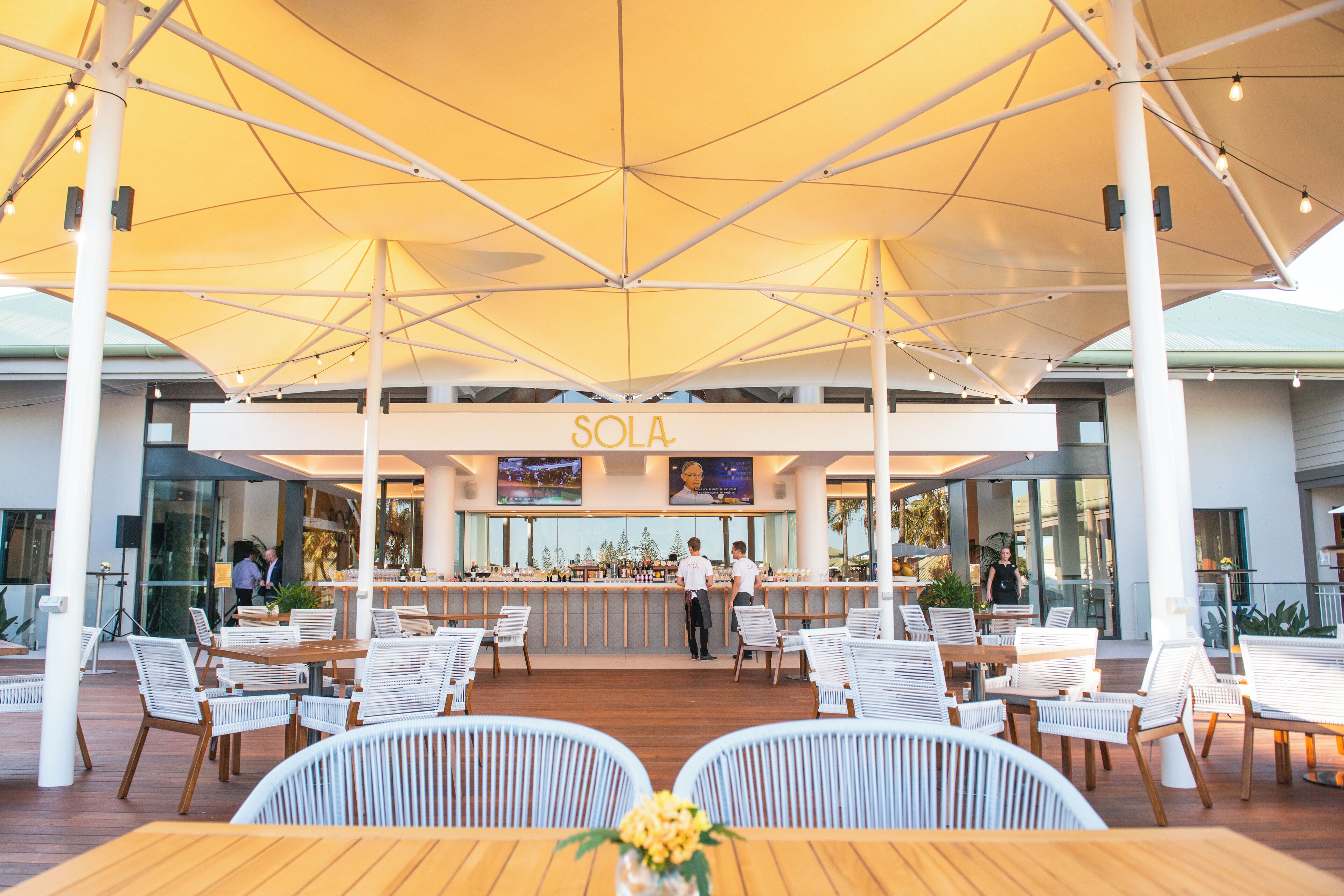 An outdoor dining area with white chairs and tables under a large canopy. A bar with a counter and stools is visible in the background.