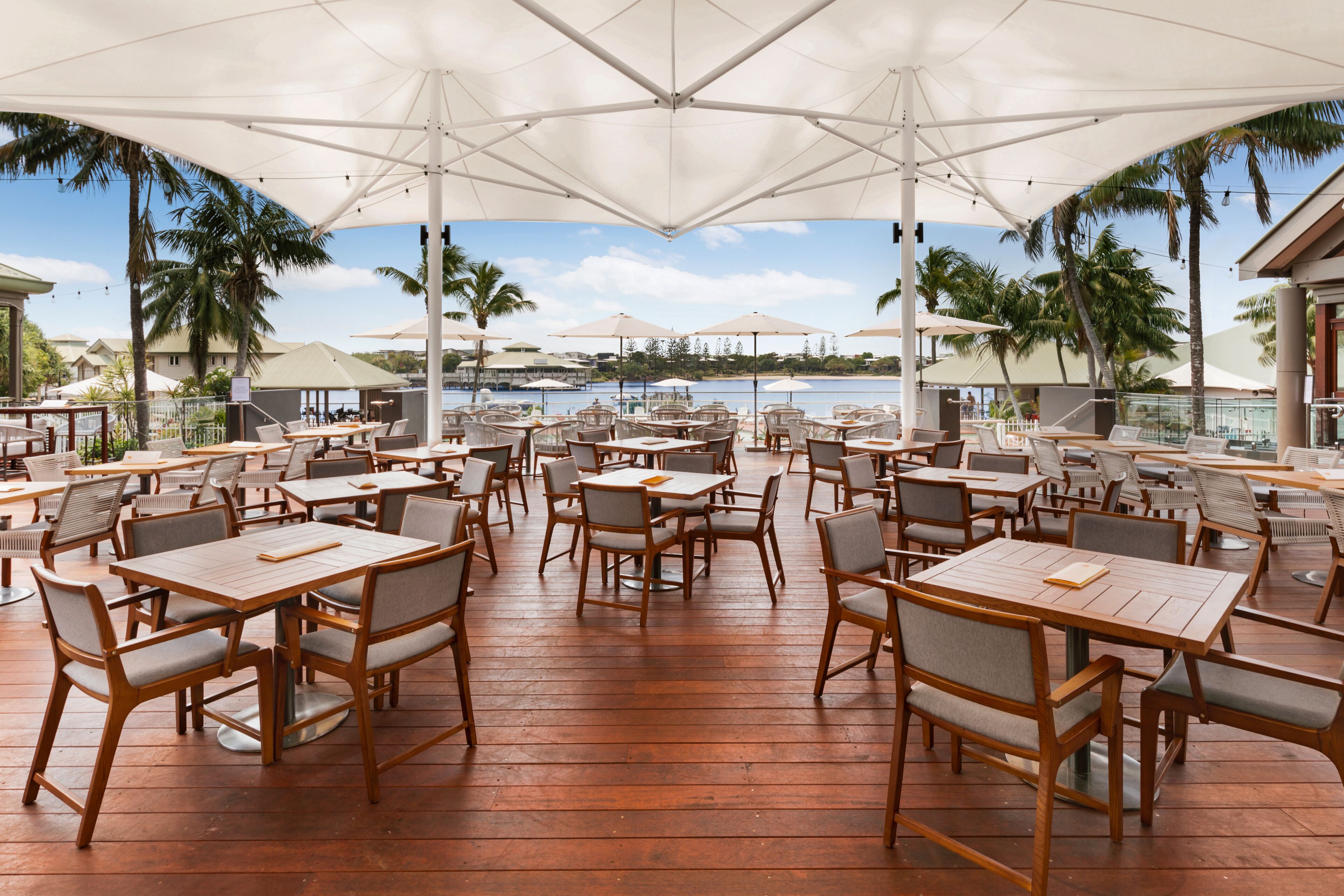 Un espace extérieur aménagé pour les repas, avec des tables et des chaises en bois, de grands parasols et une vue sur l’océan.