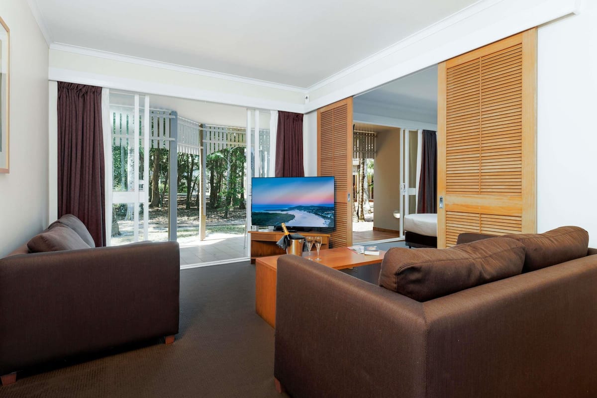 A hotel room with a large TV, two sofas, a wooden sideboard, and a sliding glass door leading to a balcony with a view of trees and a beach.
