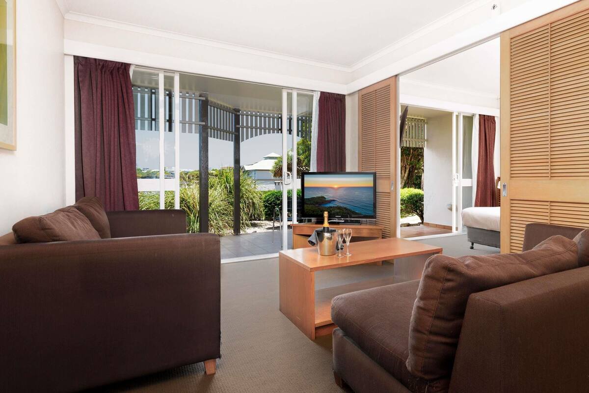 A modern living room with a flat-screen TV, brown sofas, a wooden coffee table, and a view of a balcony with a pool and greenery.