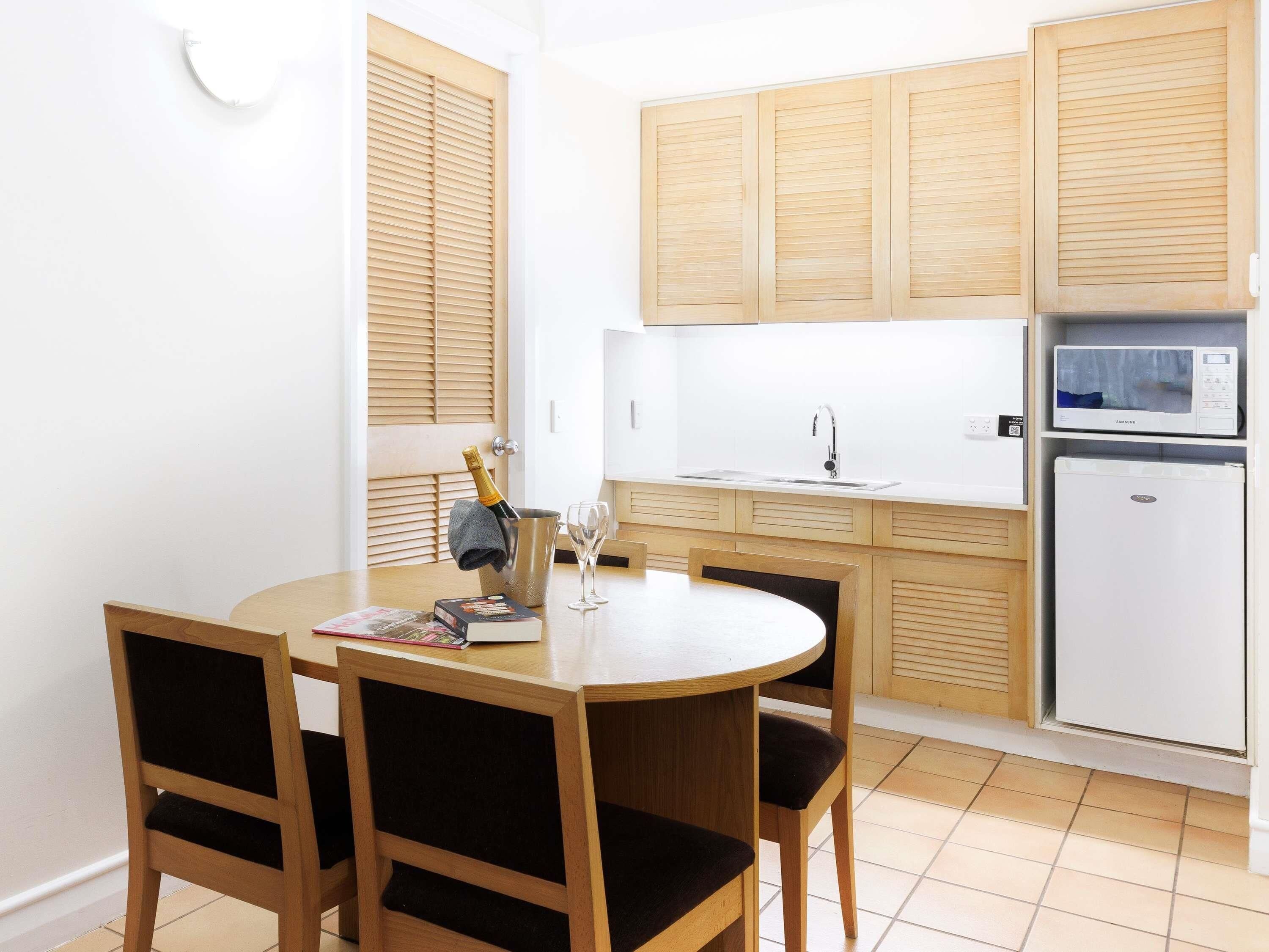 A compact kitchen with wooden cabinets, a white sink, a microwave, and a refrigerator. There is a round dining table with chairs and a bottle of champagne.