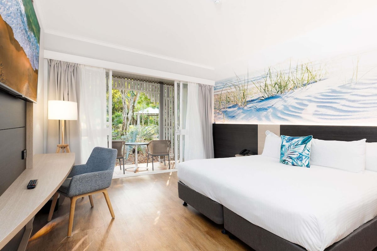A hotel room with a bed, a desk, a chair, a TV, and a view of a garden.