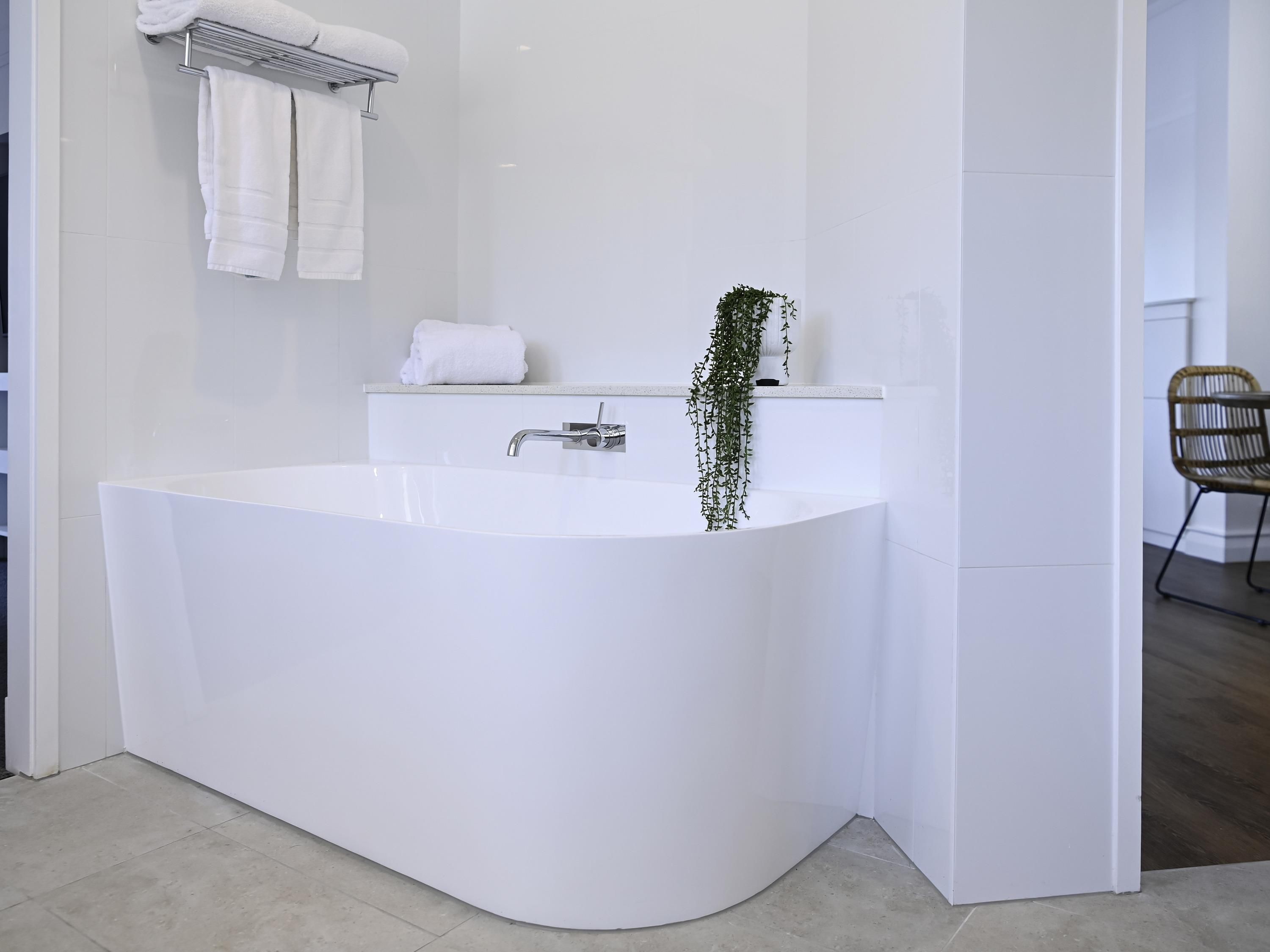 A modern bathroom with a freestanding bathtub, a towel rack with towels, a small shelf with a plant, and a wicker chair in the corner.