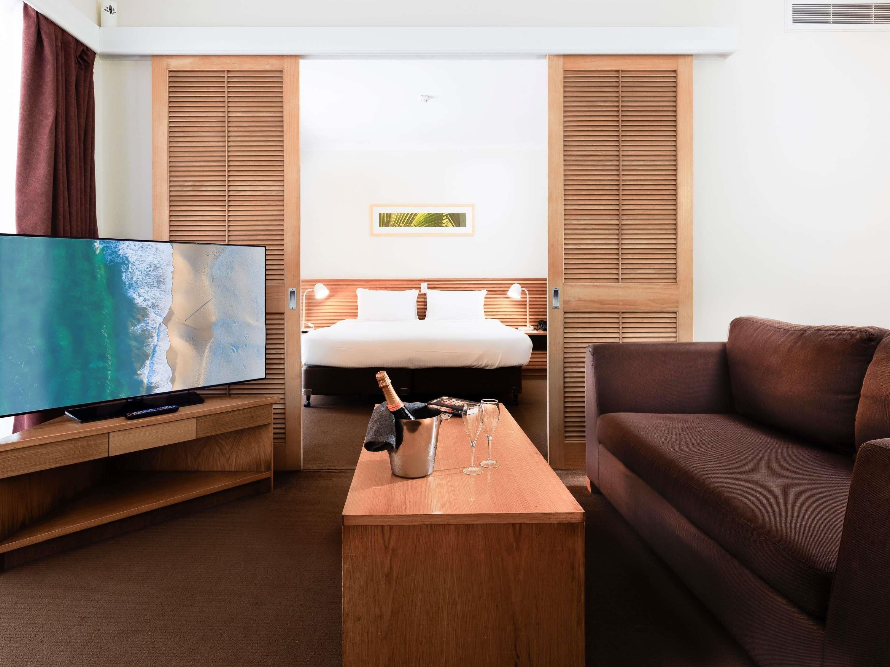 Habitació d'hotel amb un llit, un televisor, una taula de cafè de fusta amb una ampolla de xampany i gots, i un sofà marró.