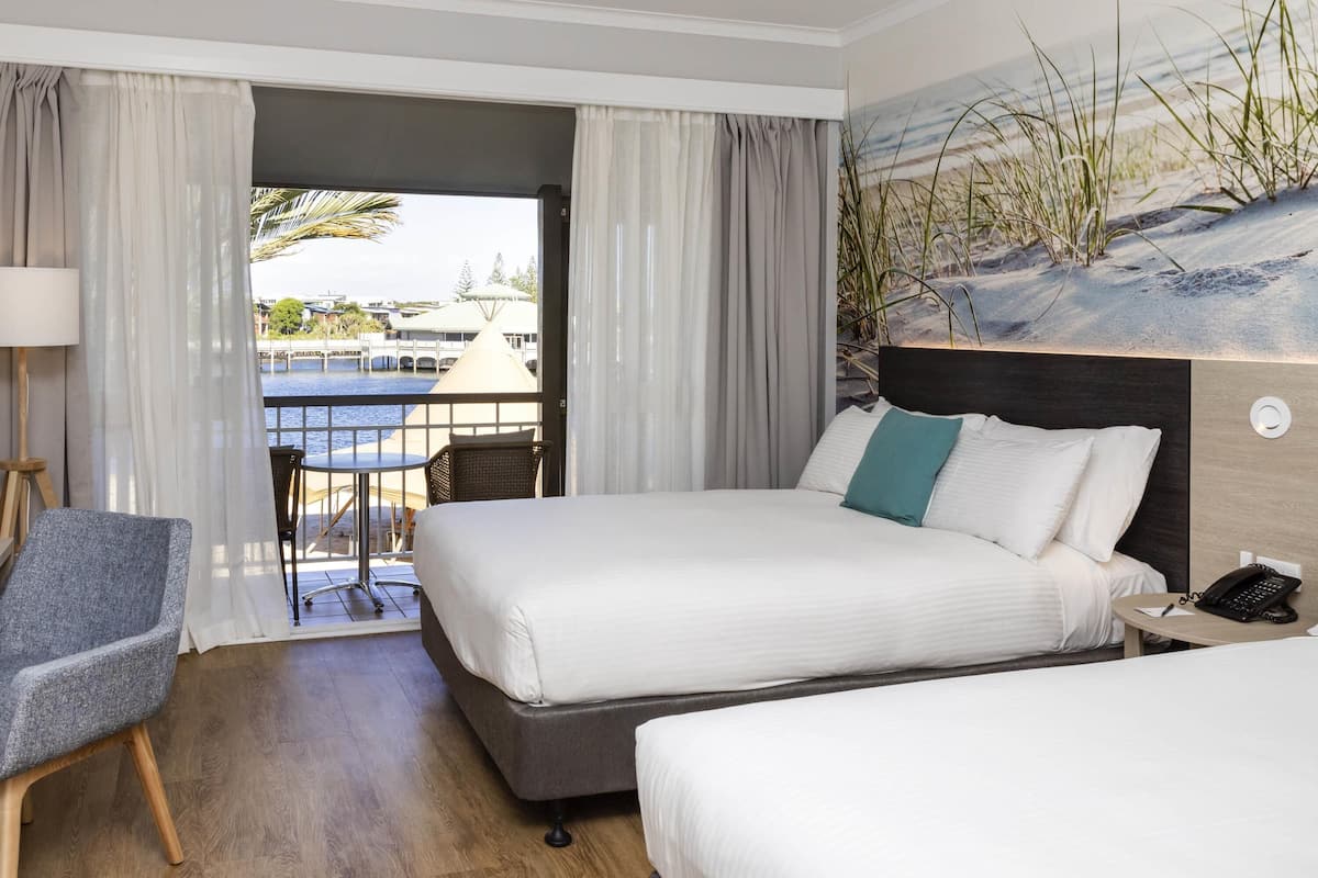 A hotel room with two beds, a chair, a table, and a view of the outside.