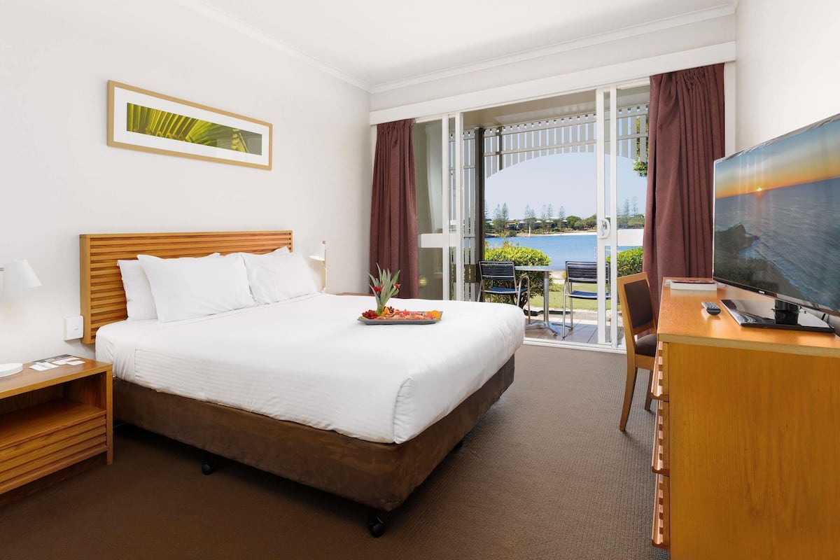 A hotel room with a bed, a desk with a TV, a dining area with chairs, and a view of the sea.