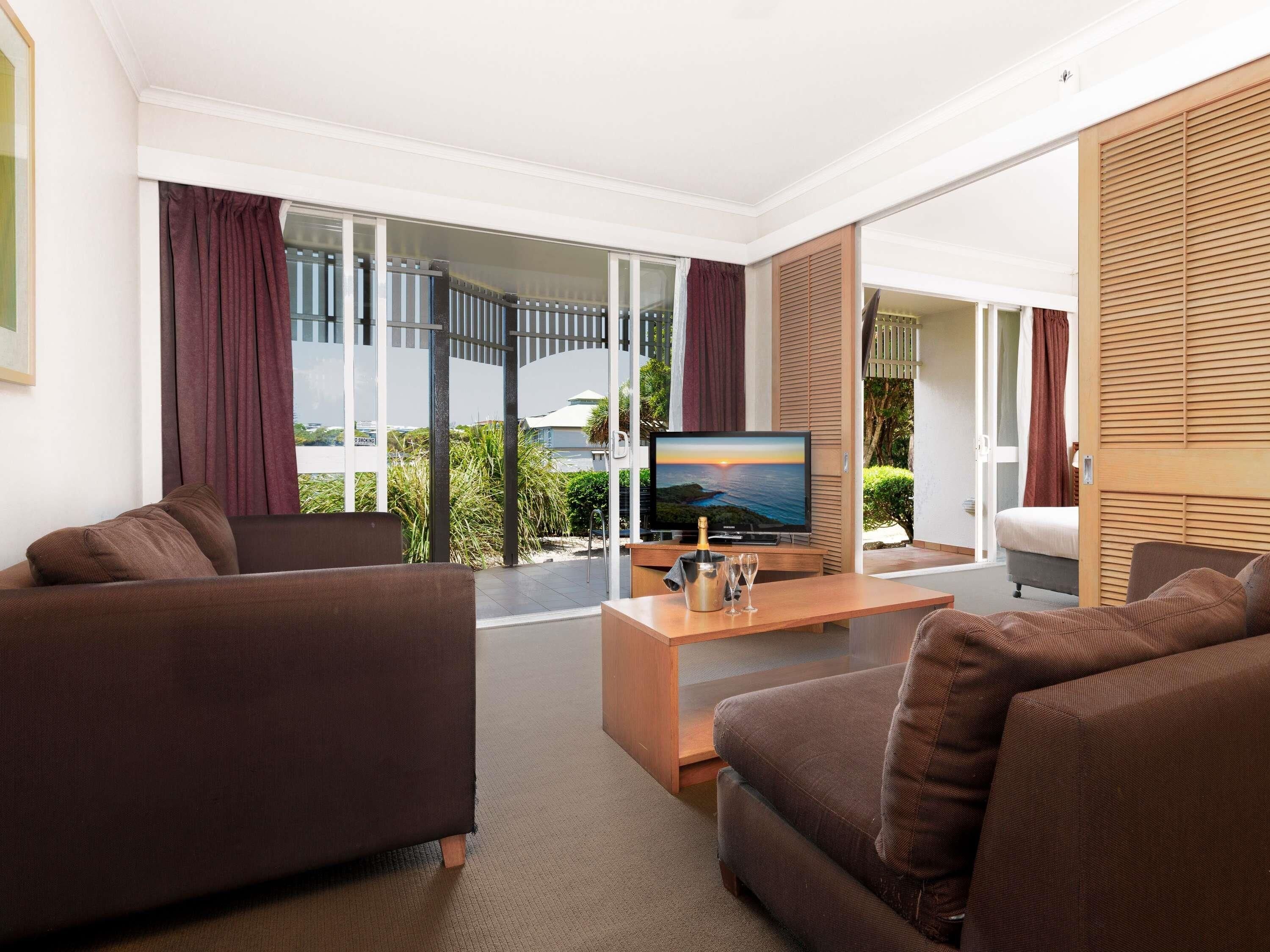 Uma sala de estar moderna com televisão de tela plana, sofás marrons, mesa de centro de madeira e vista para uma varanda com piscina e vegetação.