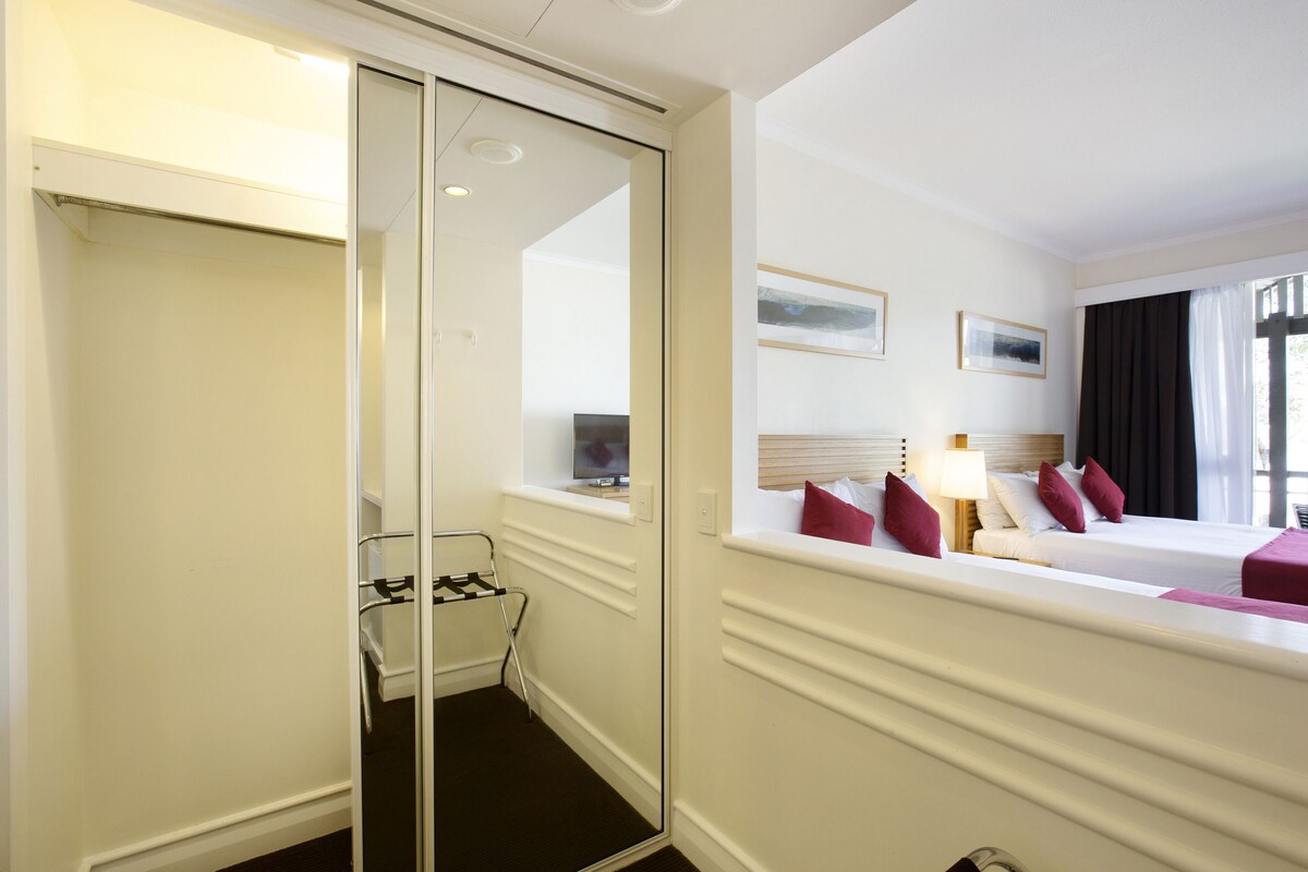 Habitación de hotel con un espejo grande, una cama con almohadones rojos, una mesita de noche con una lámpara y una ventana con cortinas.