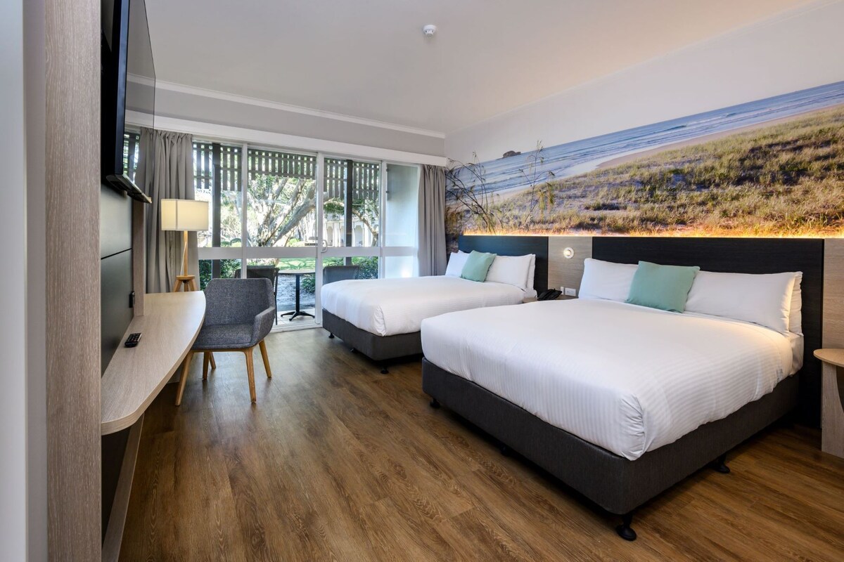 Habitación de hotel con dos camas, un escritorio, una silla y un ventanal con vistas a la playa.