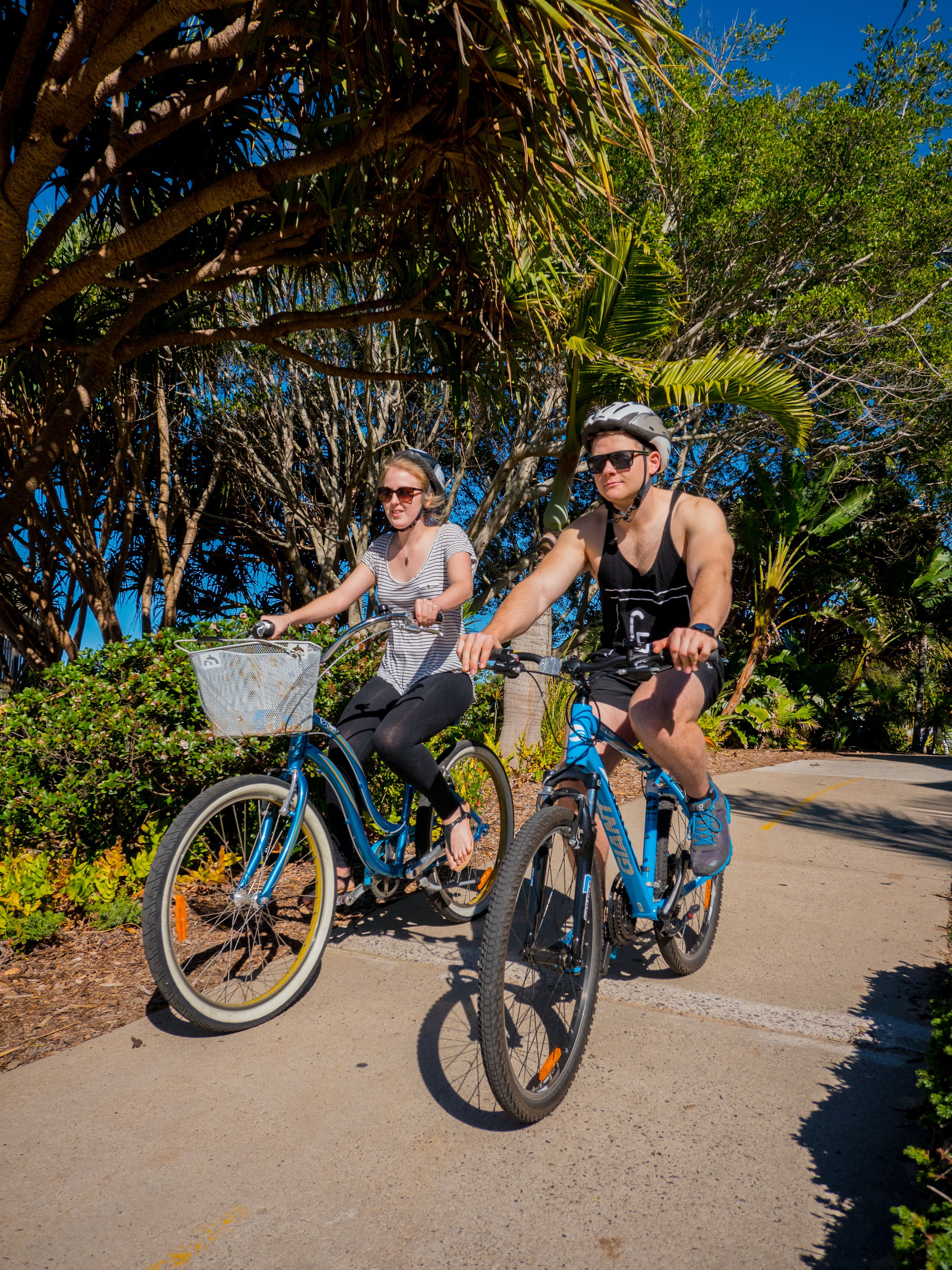 Two people riding bicycles on a paved path surrounded by lush greenery and trees.