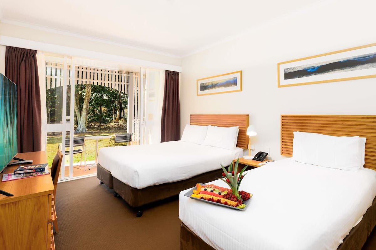 A hotel room with two beds, a desk, and a balcony with a view of trees.