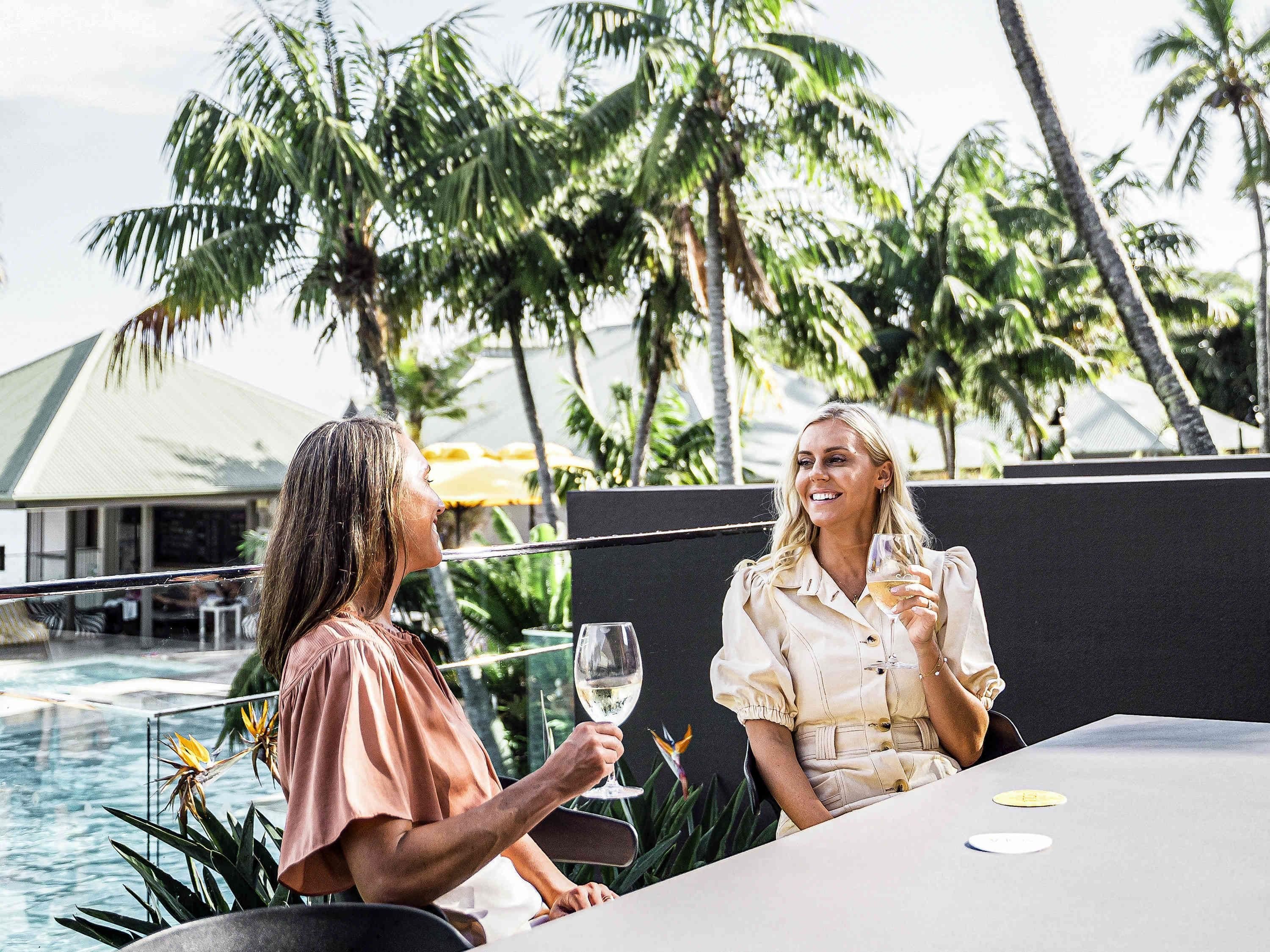 Two people sitting at a table by a pool with palm trees in the background.