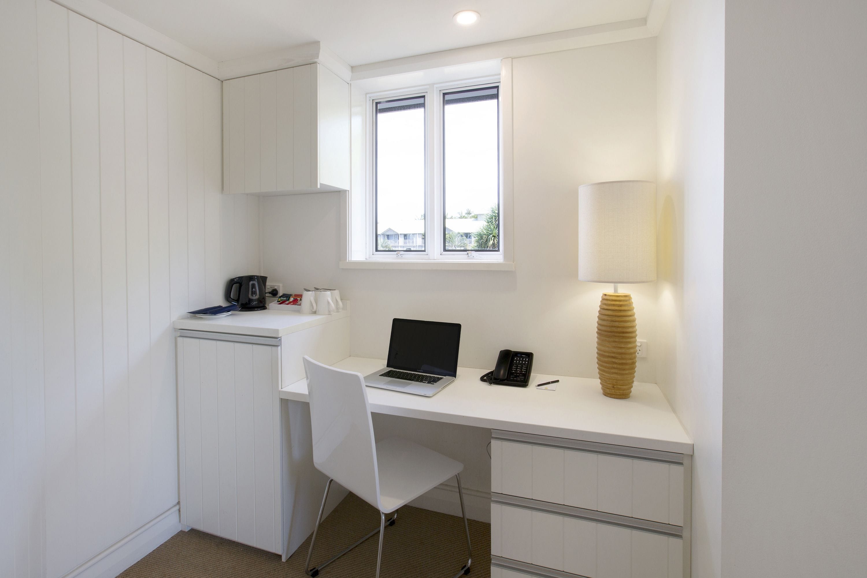 A compact, white room with a desk, a laptop, a telephone, a lamp, and a window.