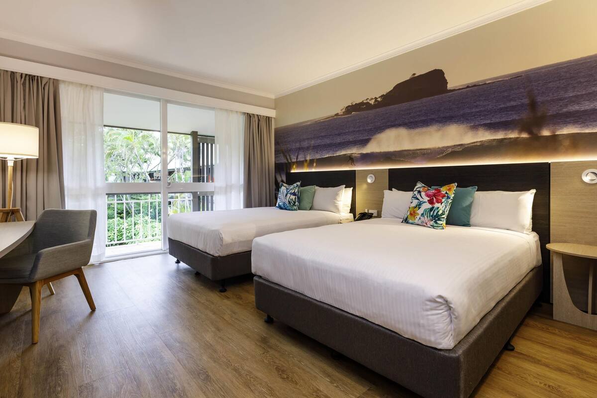 A hotel room with two beds, a desk, and a balcony with a view of the ocean.