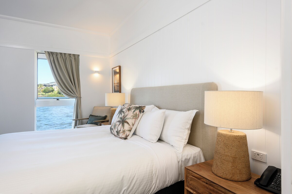 Habitación de hotel con una cama grande, una lámpara de noche, una silla y vista al mar.
