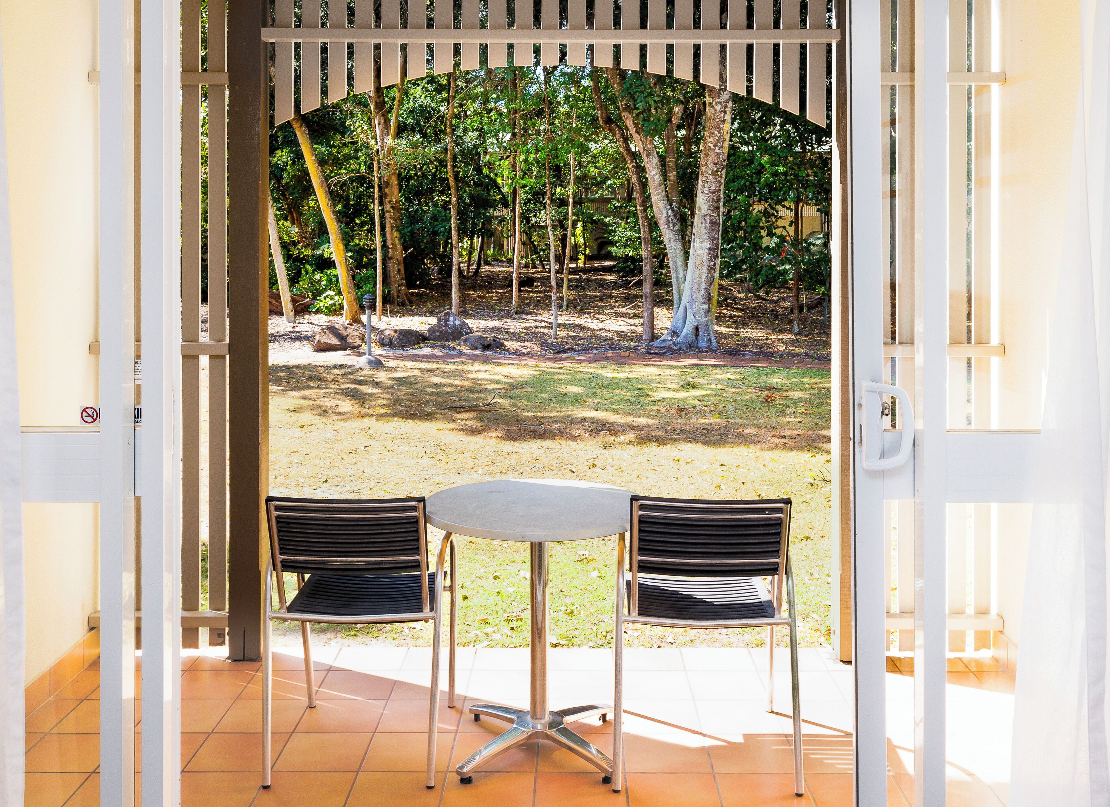 An open patio with two chairs and a table, a view of a wooded area through a white fence, and a tiled floor.