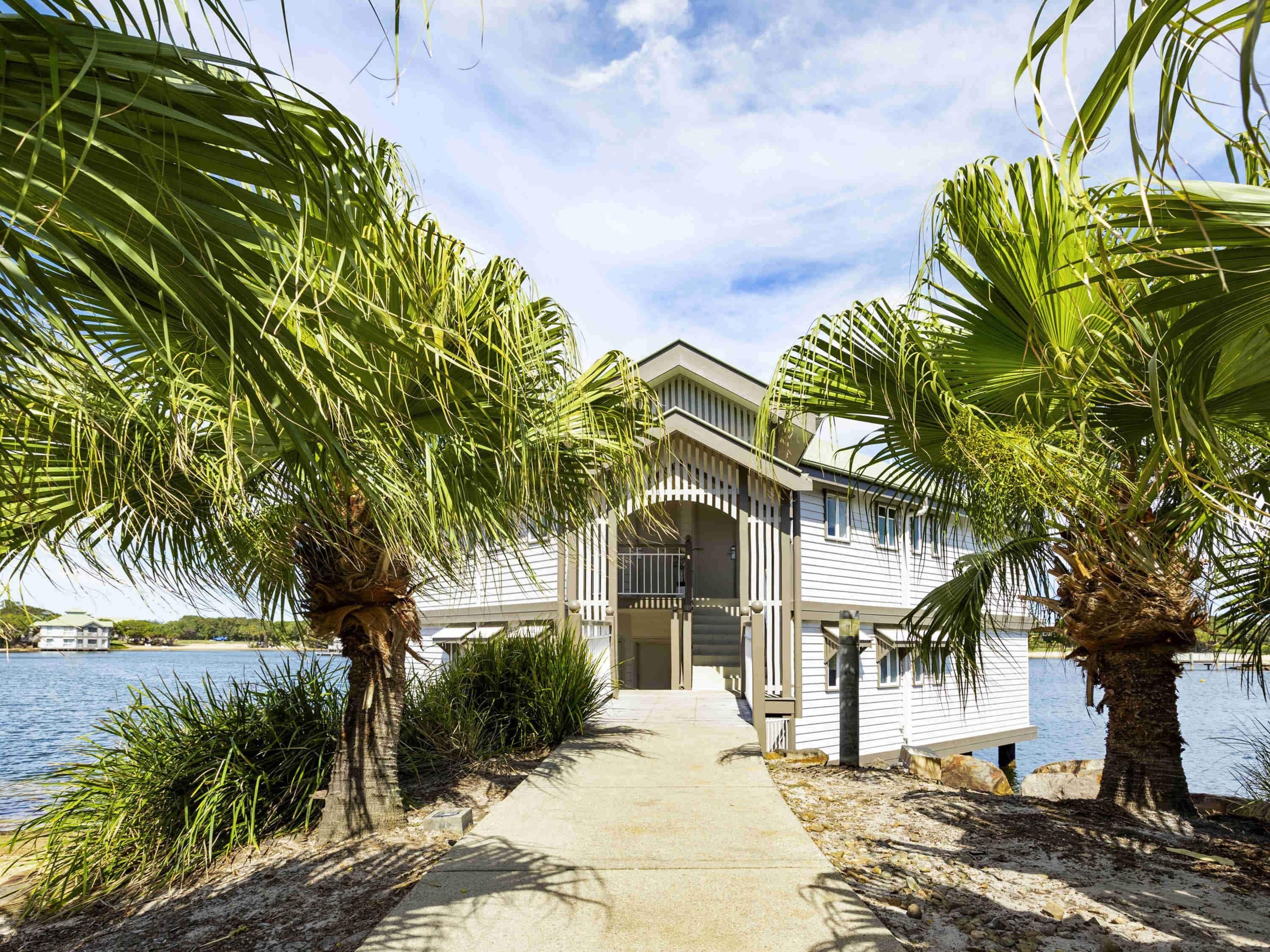 A waterfront property with a pathway leading to a building, surrounded by palm trees and a body of water.