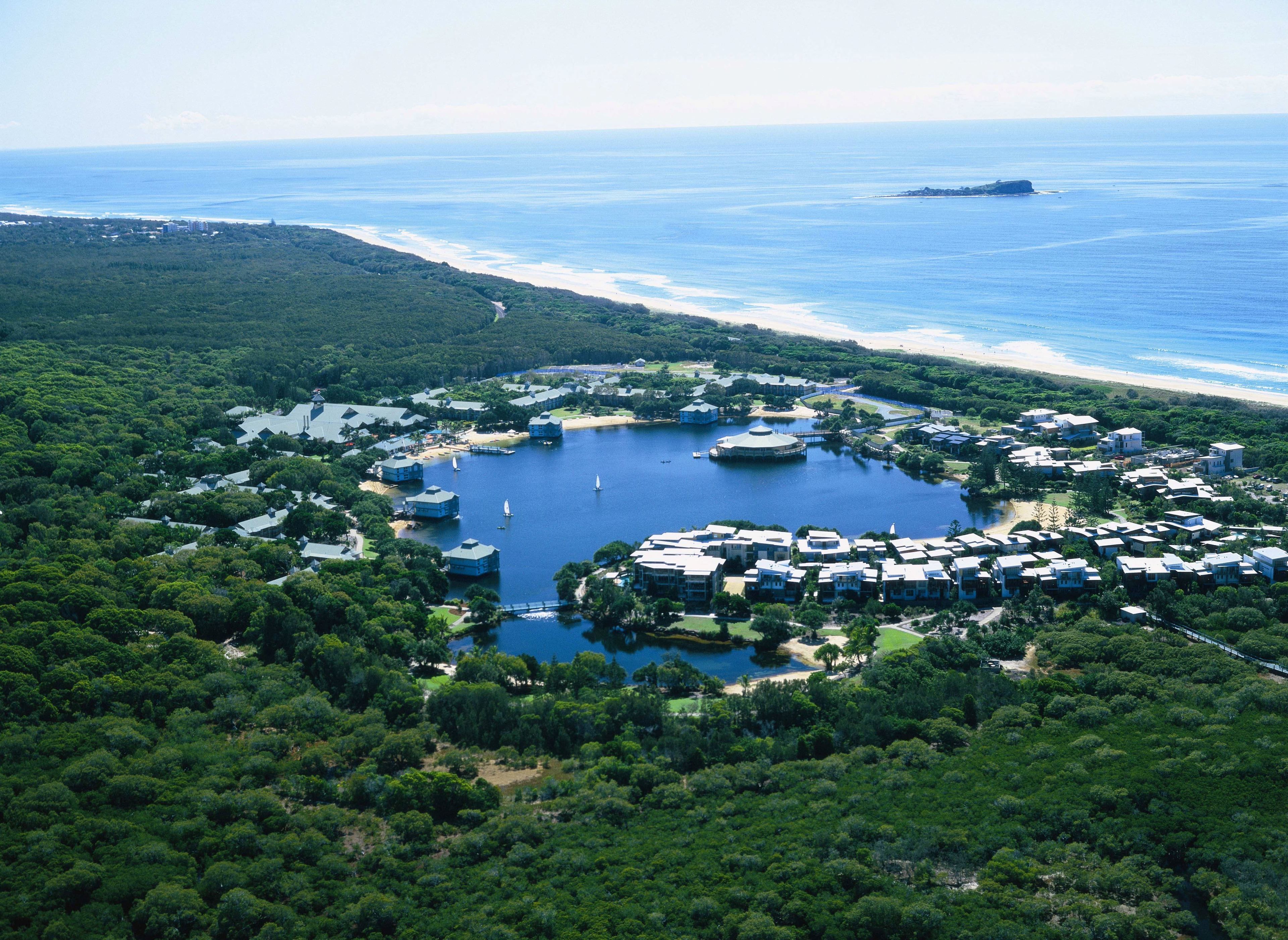 An aerial view of a coastal resort with multiple buildings, a central lake, and a beach.