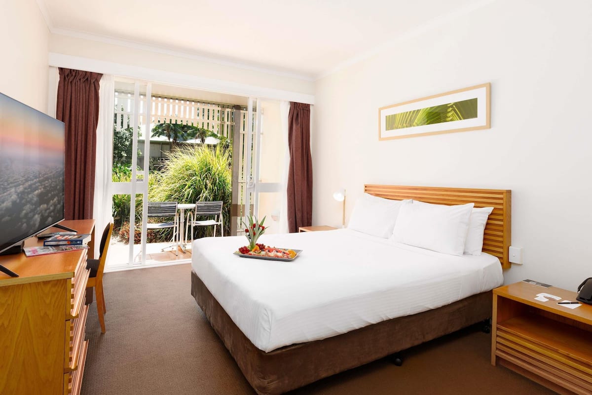 A hotel room with a bed, a TV, a desk with a computer, a dining table, and a view of greenery outside.
