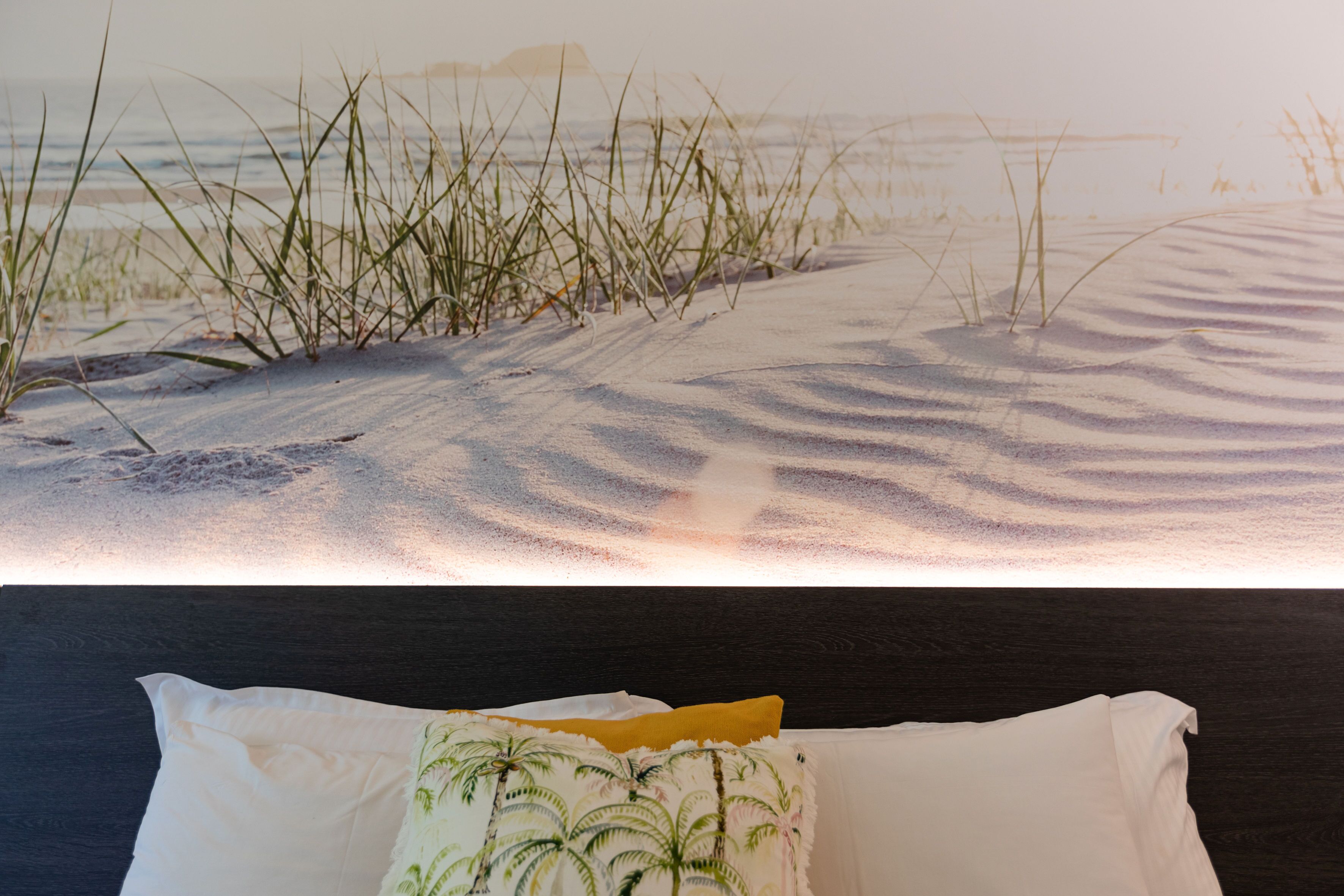 Uma cama com travesseiro com cena de praia, cabeceira escura e vista para uma praia arenosa com grama.