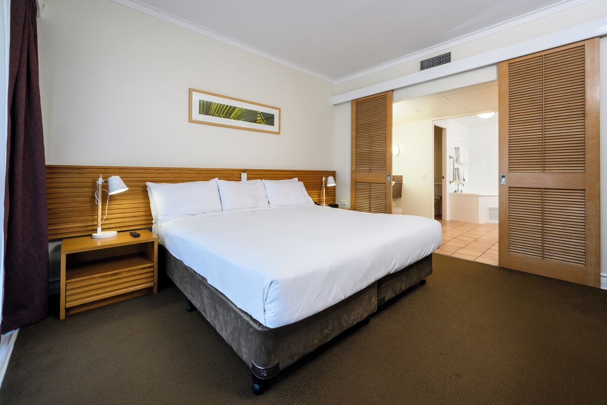 Una cama bien hecha en una habitación de hotel, con cabecera de madera, una mesita de noche con lámpara y un cuadro en la pared.