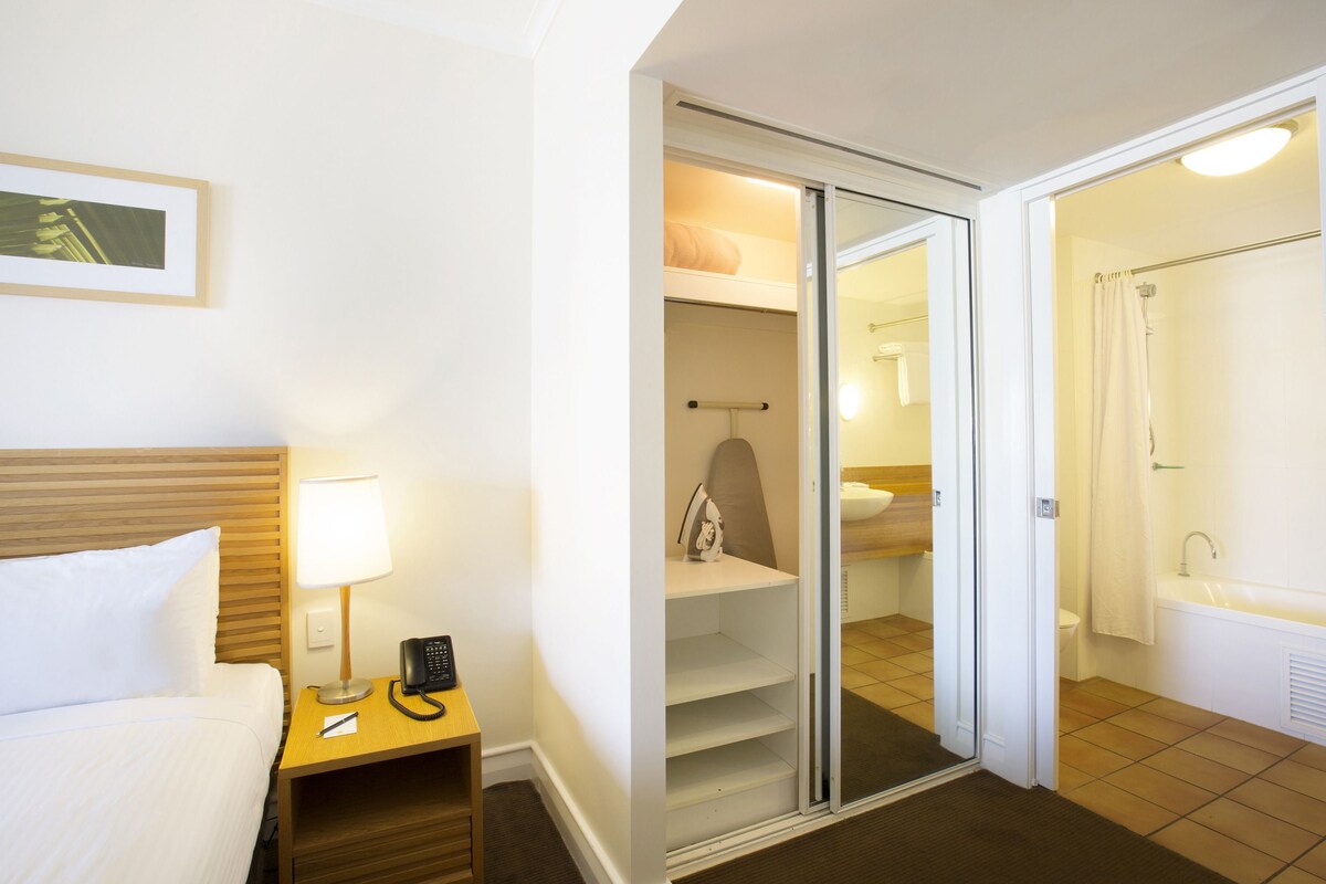 Habitación de hotel con cama, mesita de noche con lámpara y teléfono, armario con puertas correderas y baño con bañera e inodoro.