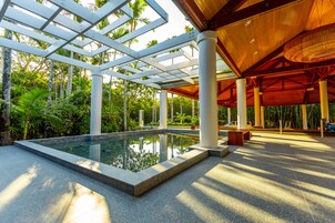 Zona de piscina con cerramiento de vidrio, rodeada de palmeras y una pérgola de madera.