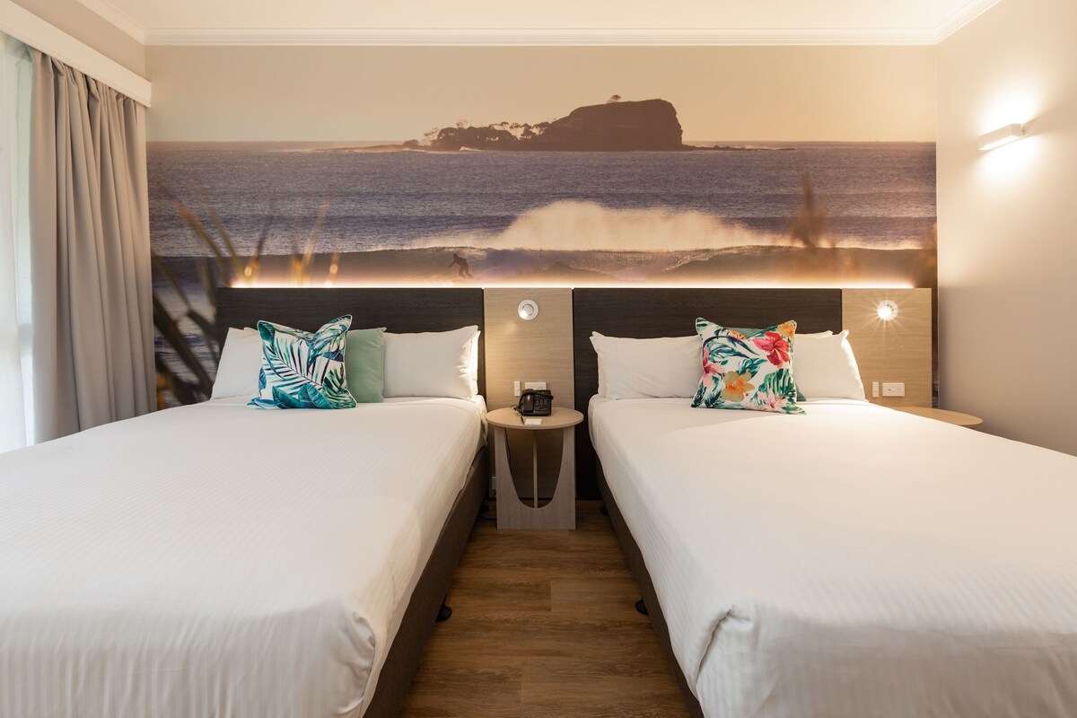 Una habitación de hotel con dos camas, una mesita de noche y un mural grande con una playa.