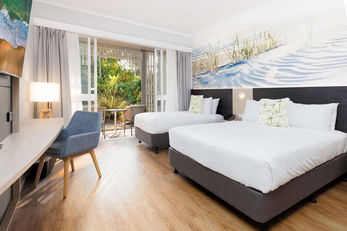 A hotel room with two beds, a desk, and a chair. There is a view of greenery through the window.