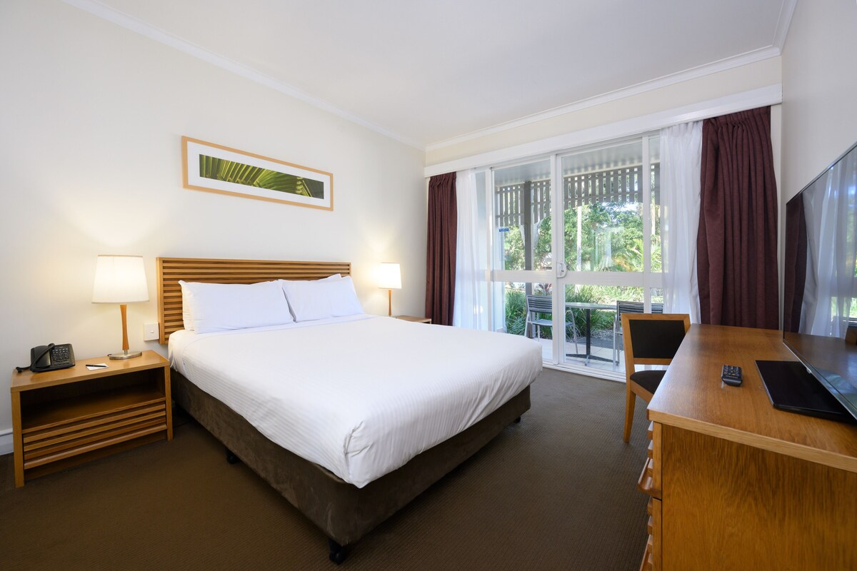 Una habitación de hotel con cama, mesitas de noche, escritorio, silla, televisión y ventana con cortinas.