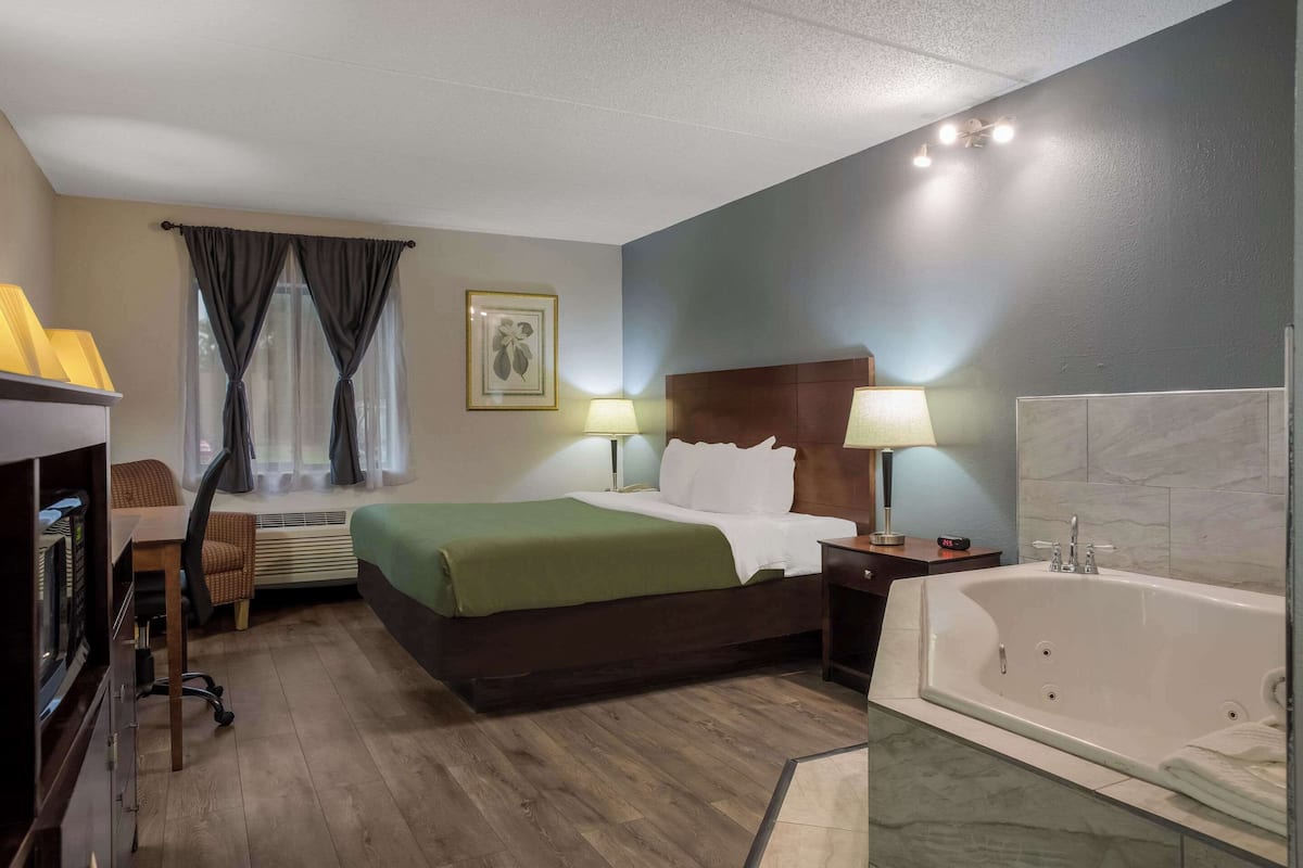 A hotel room with a bed, a desk, a chair, a window with curtains, a wall-mounted picture, a nightstand with a lamp, and a bathtub.